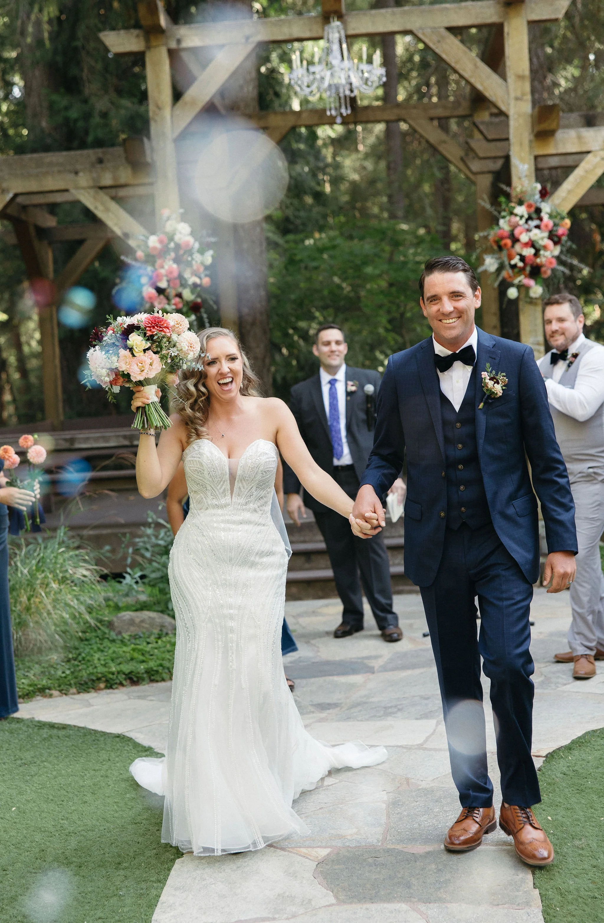 A new bride and groom smile and walk hand-in-hand after their wedding ceremony in an outdoor setting with wooden structures and hanging chandeliers, surrounded by friends and family, with the bride holding a colorful bouquet.