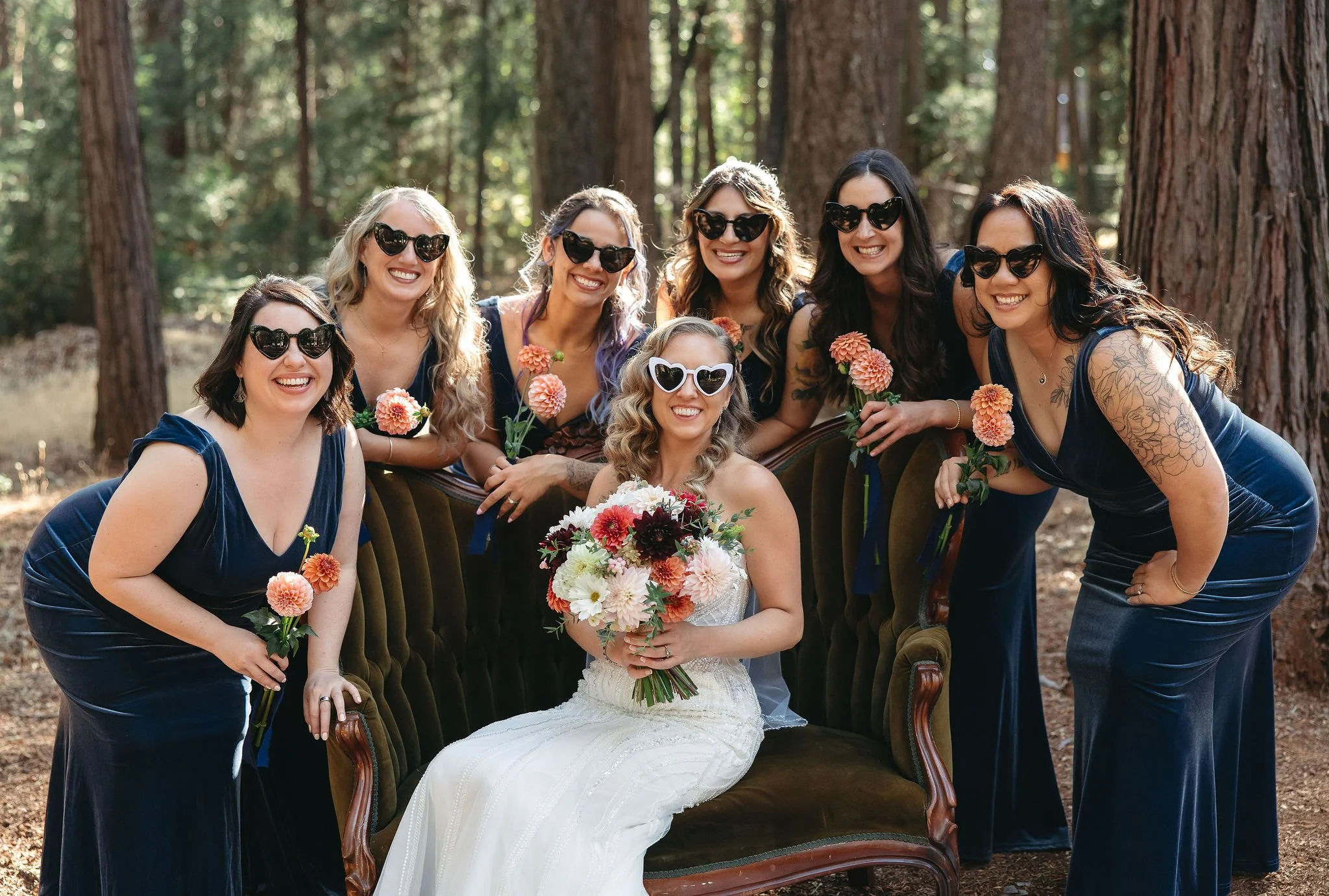 A bride in a white dress holding a bouquet of flowers, surrounded by seven women in matching navy velvet dresses and sunglasses, in a forest setting.