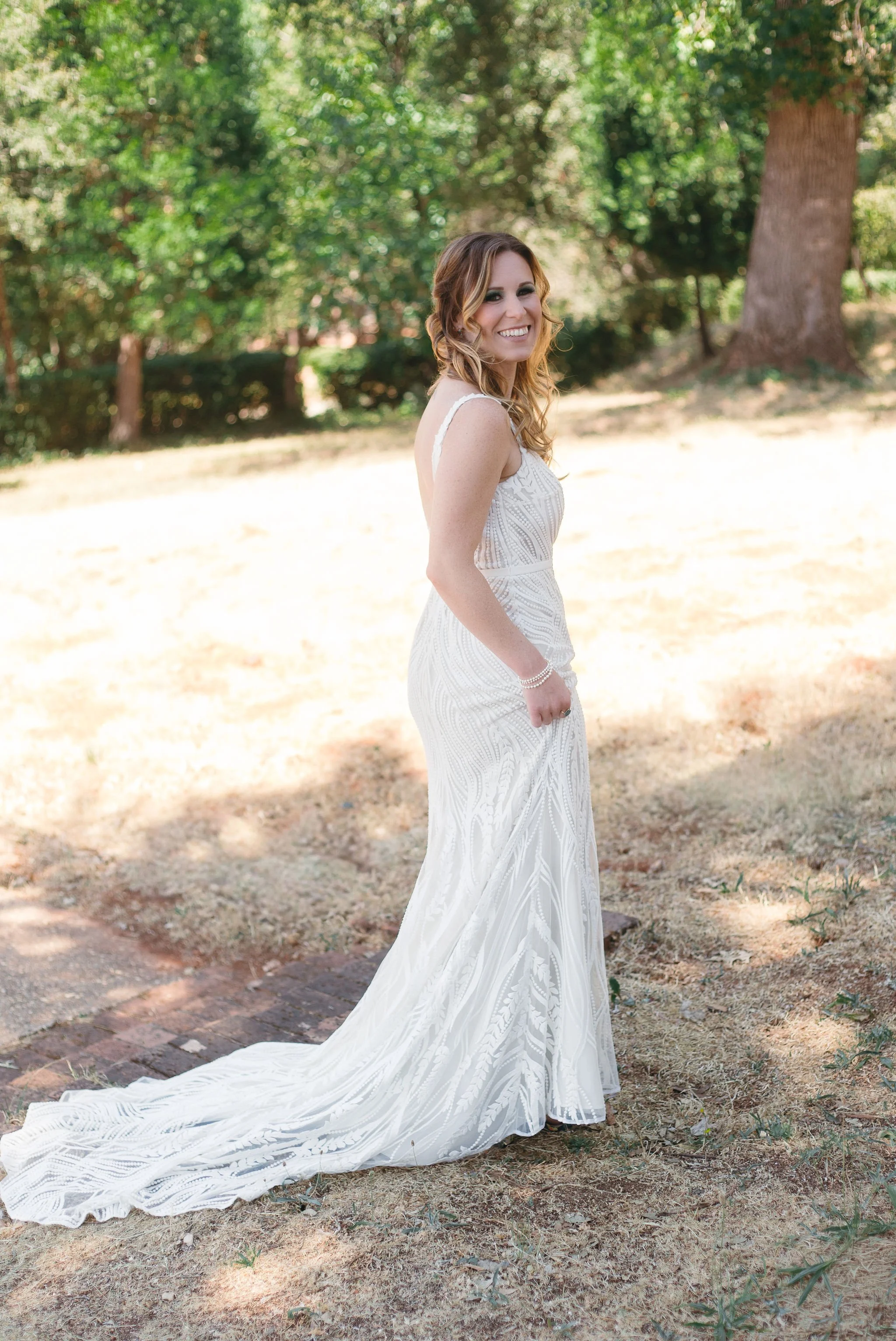 A woman in a white wedding dress standing outdoors on grass, smiling and looking at the camera, with trees and sunlight in the background.