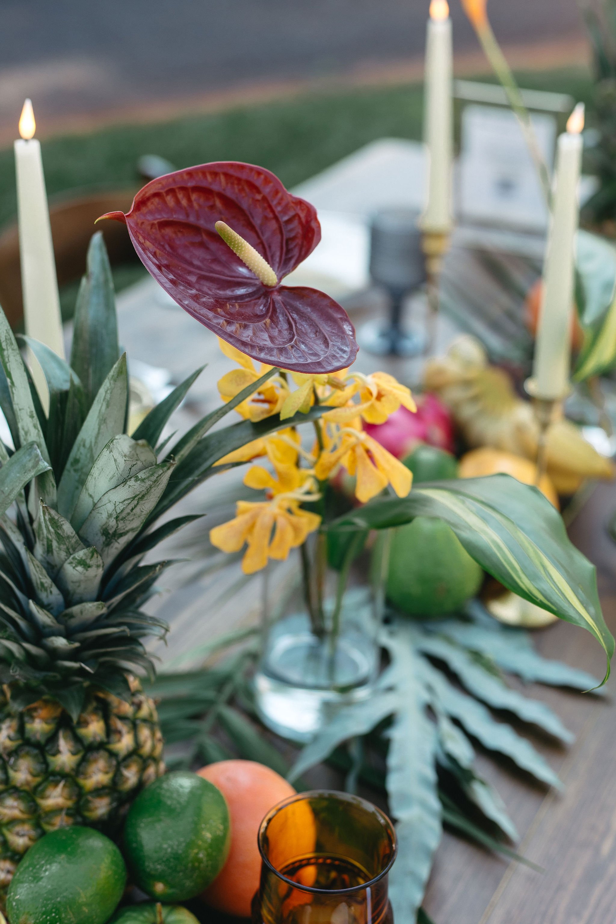 Tropical fruit and floral centerpiece with candles on a table and an outdoor background.
