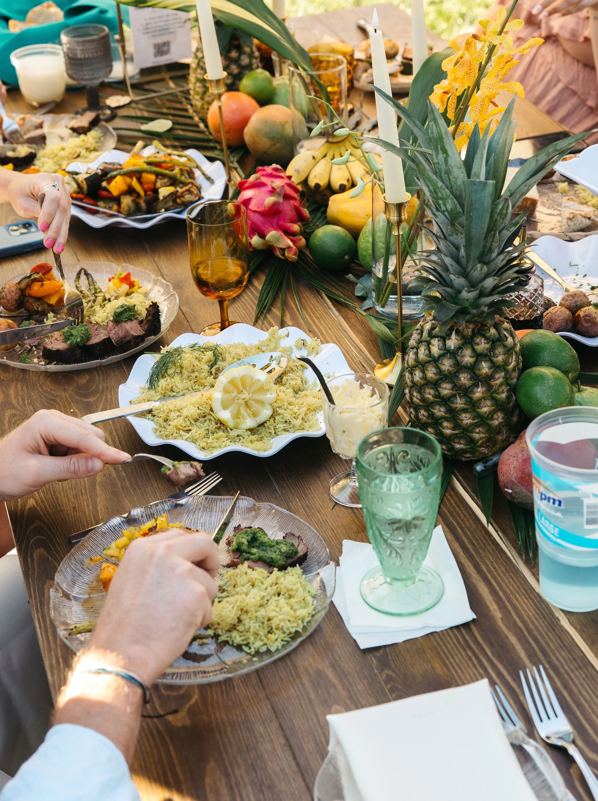 A festive outdoor meal with tropical fruits, candles, and dishes including rice, grilled meat, and vegetables on a wooden table.