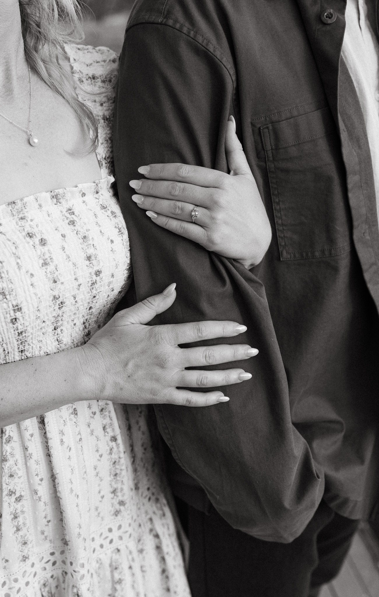 A woman touching a man's arm with her left hand, showing an engagement ring, and her right hand resting on his chest. Both are partially visible, dressed in casual clothing, with the woman wearing a floral dress and the man in a dark shirt or jacket.