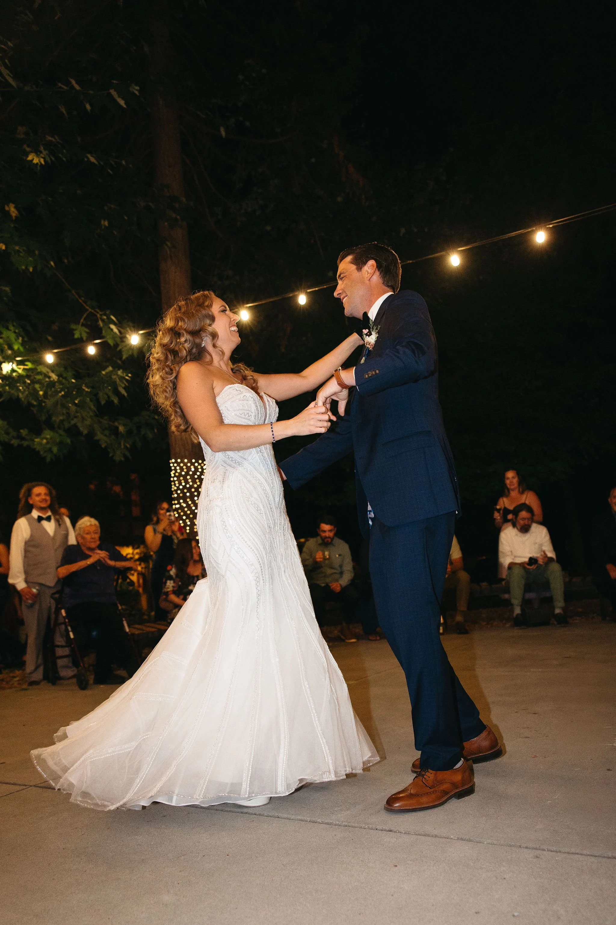 A bride and groom dancing at their wedding reception outdoors at night under string lights, with guests watching in the background.