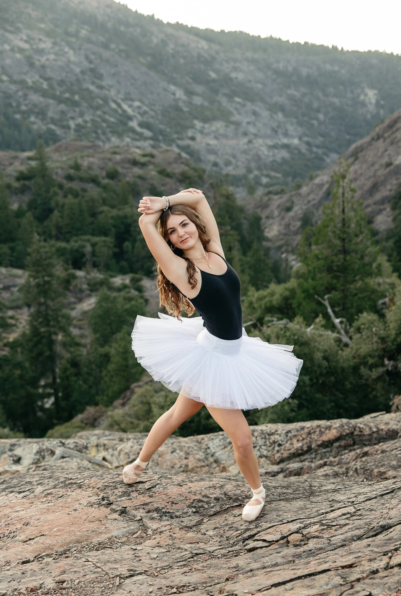 A woman in ballet attire, wearing a black leotard and a white tutu, practicing ballet on a rocky outdoor terrain with mountain and forest in the background.