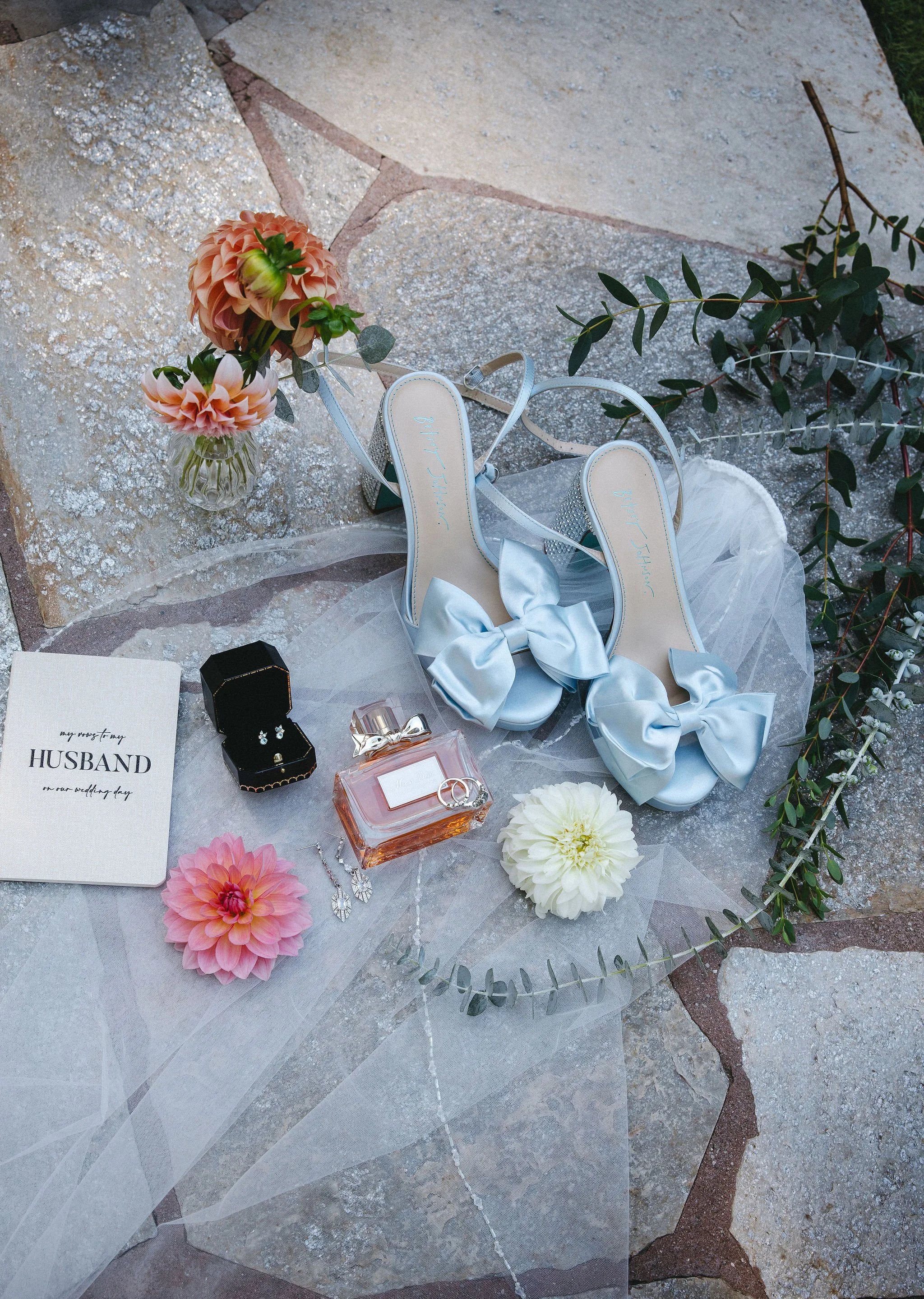 Wedding shoes, jewelry, flowers, and a wedding invitation arranged on a stone surface.
