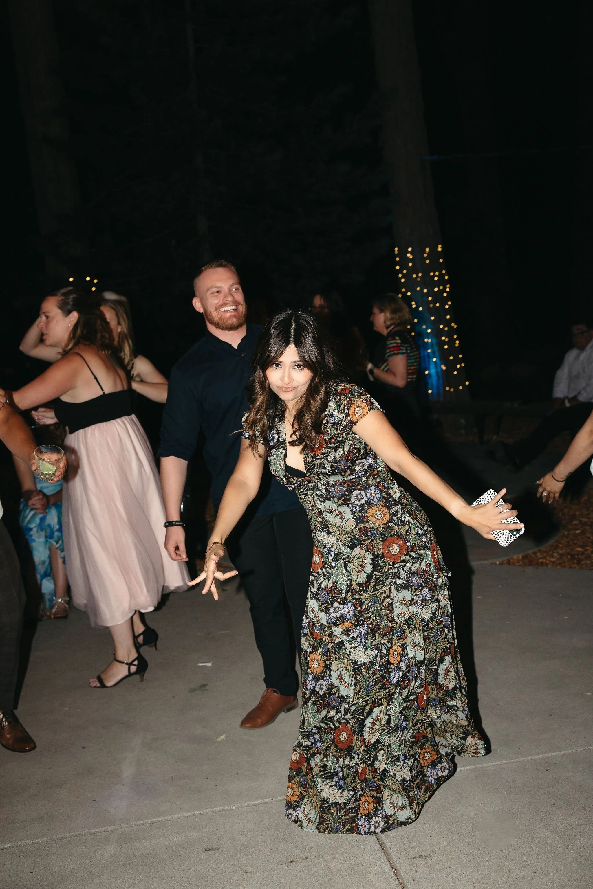 People dancing outdoors at night, some holding drinks, with a tree wrapped in string lights in the background.