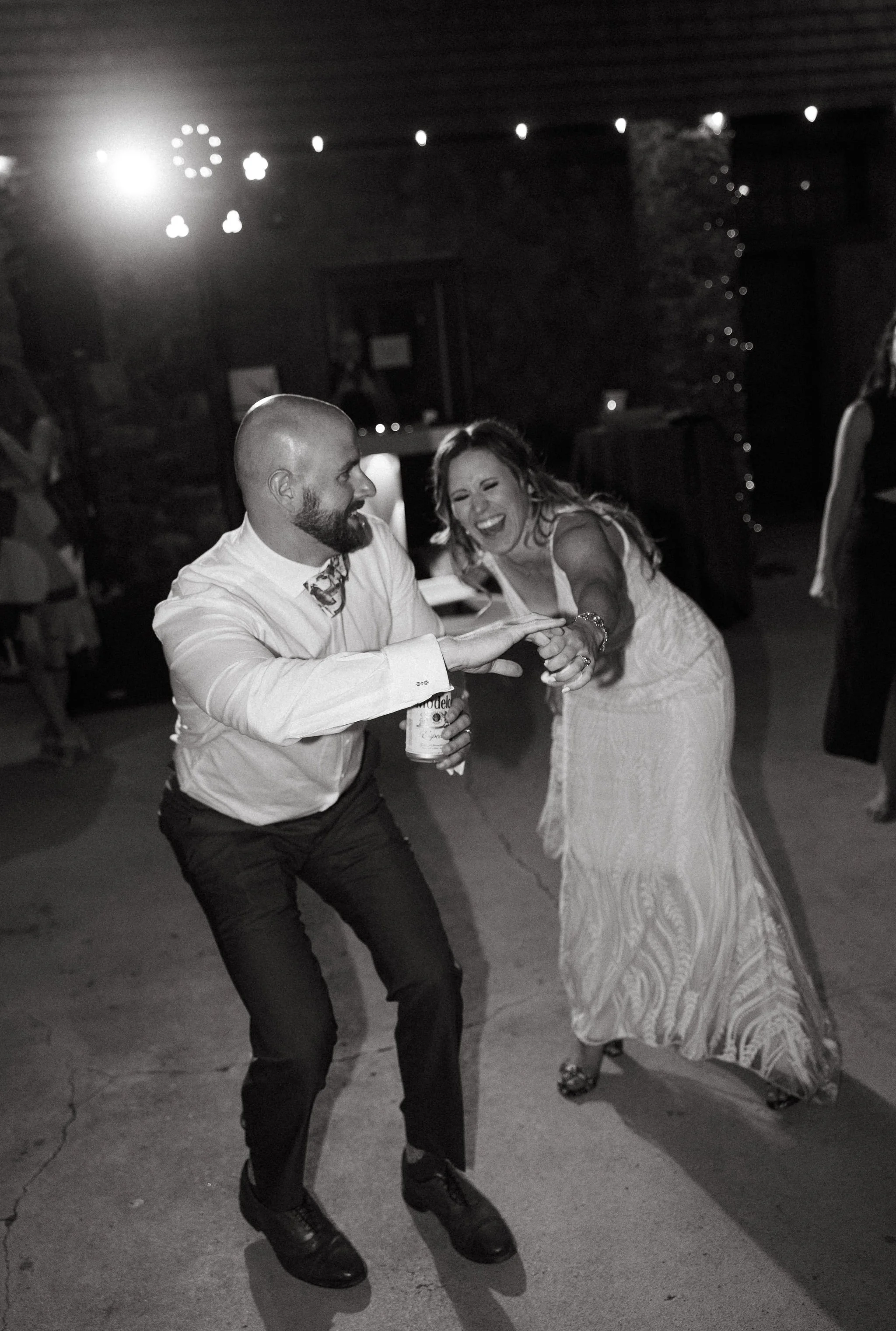 A man and woman are dancing and laughing together at a celebration or wedding reception, with others in the background.