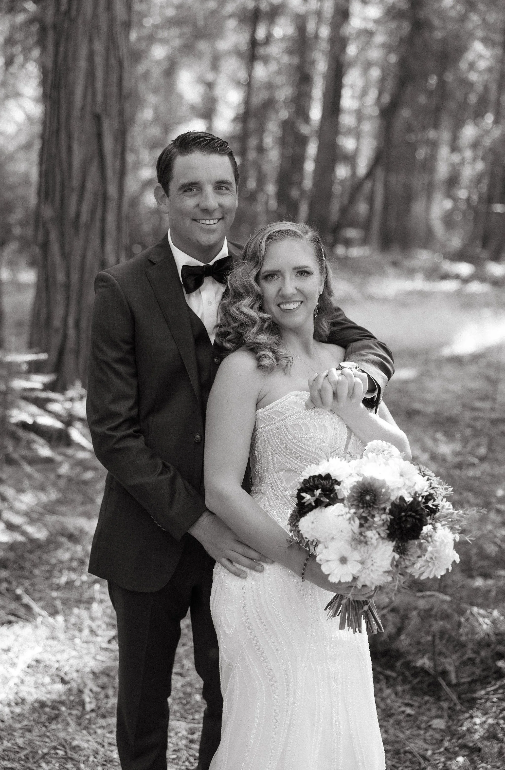 Black and white photograph of a newlywed couple in a forest. The man is wearing a suit with a bowtie, and the woman is in a wedding dress holding a bouquet of flowers. The man has his arm around the woman's shoulder, and they are smiling.