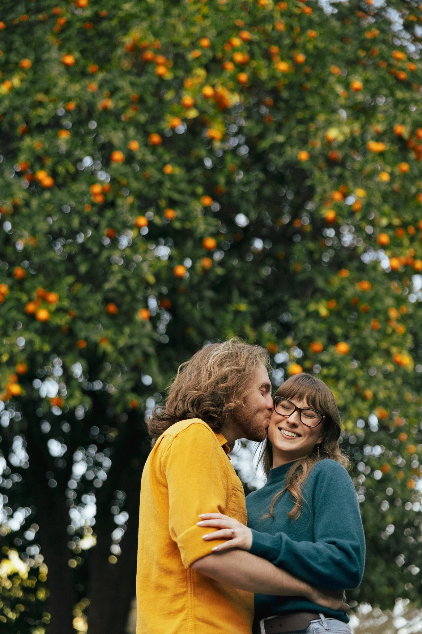 A woman and a man sharing a kiss while hugging each other, smiling, in front of a tree with orange fruit.