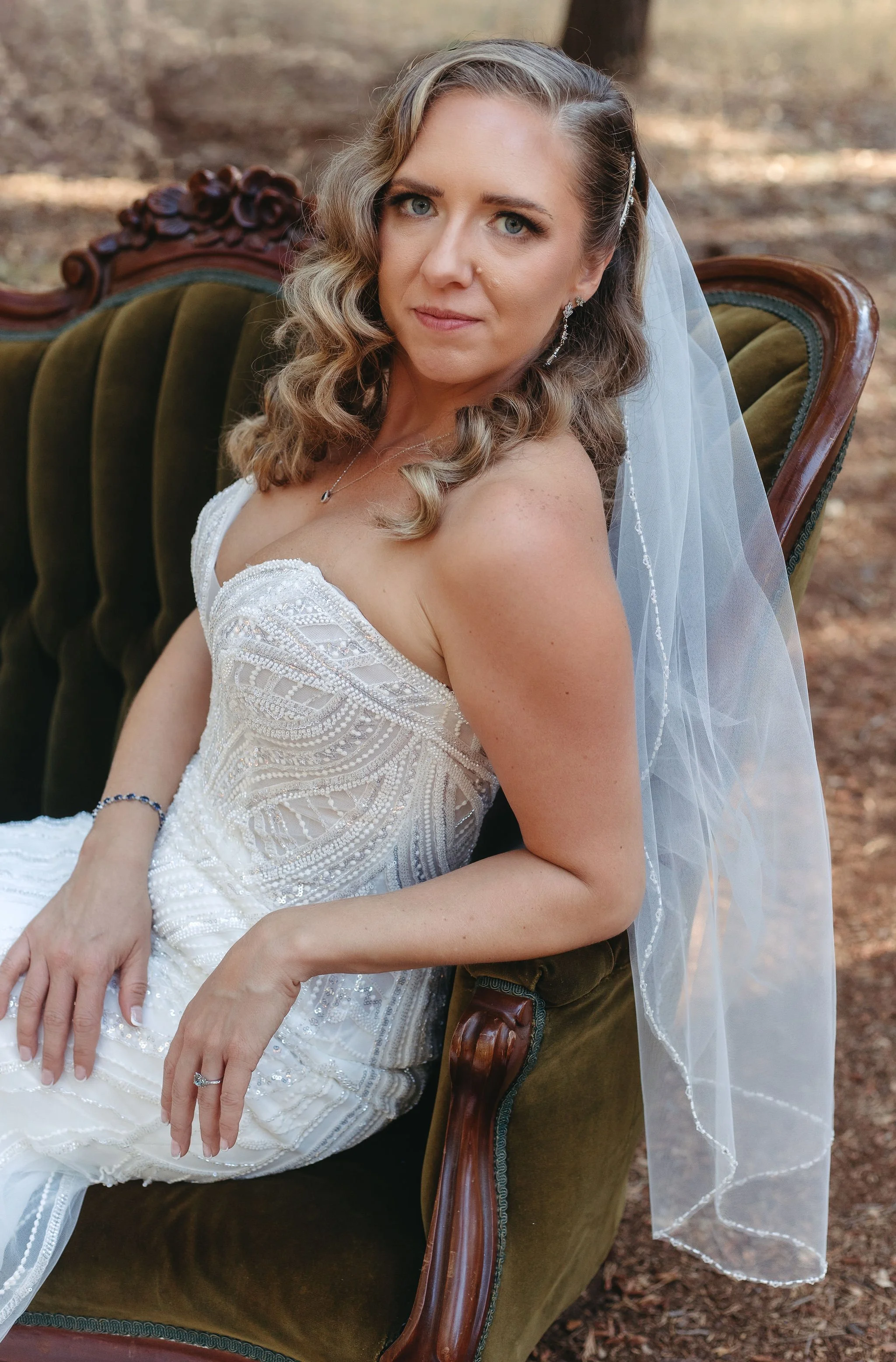 A bride with wavy blond hair and blue eyes sitting on an antique green velvet armchair outdoors, wearing a white beaded wedding dress and veil.