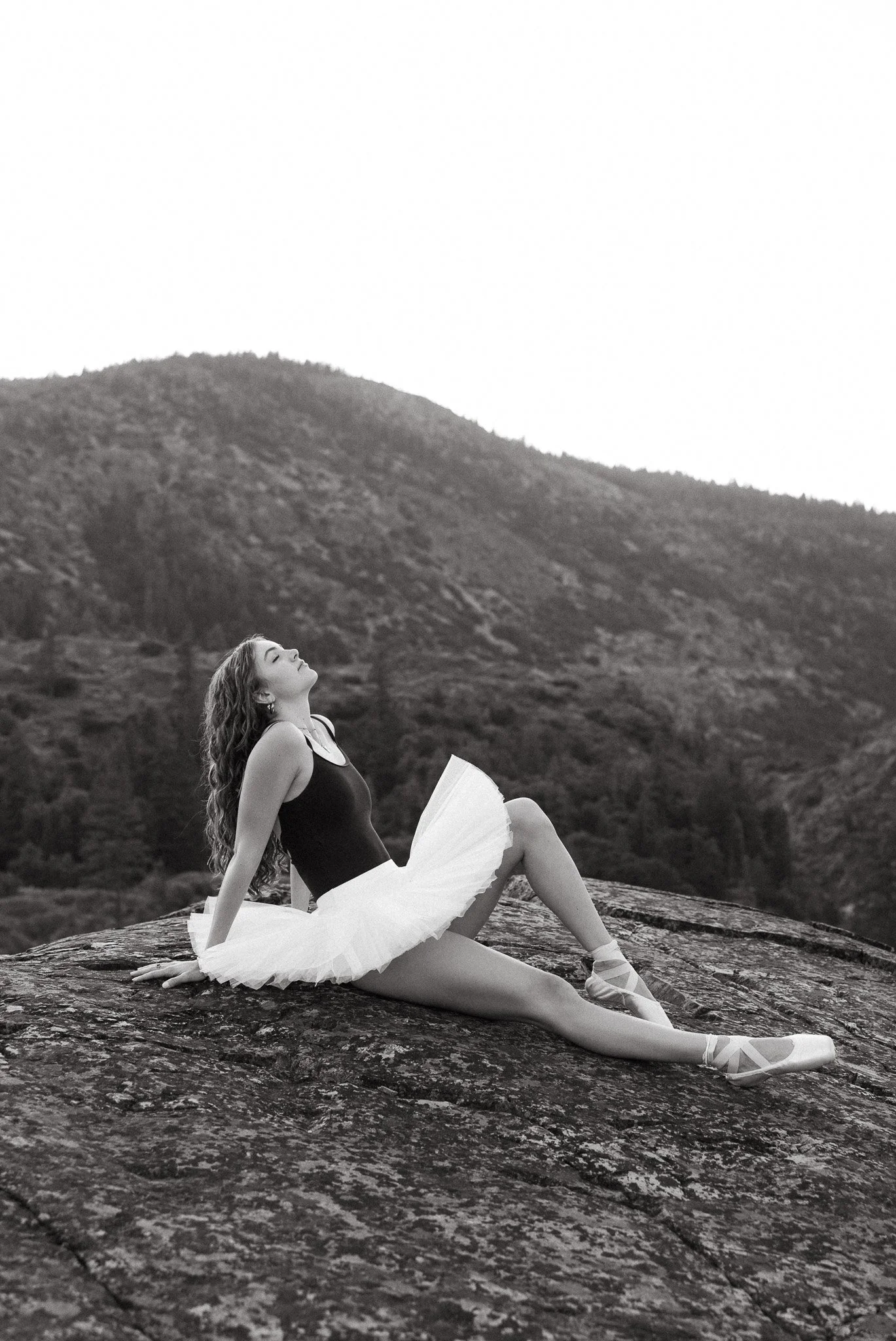 A ballet dancer sits on a rocky surface outdoors, wearing a black leotard, white tutu, and ballet slippers, with a scenic mountain background.