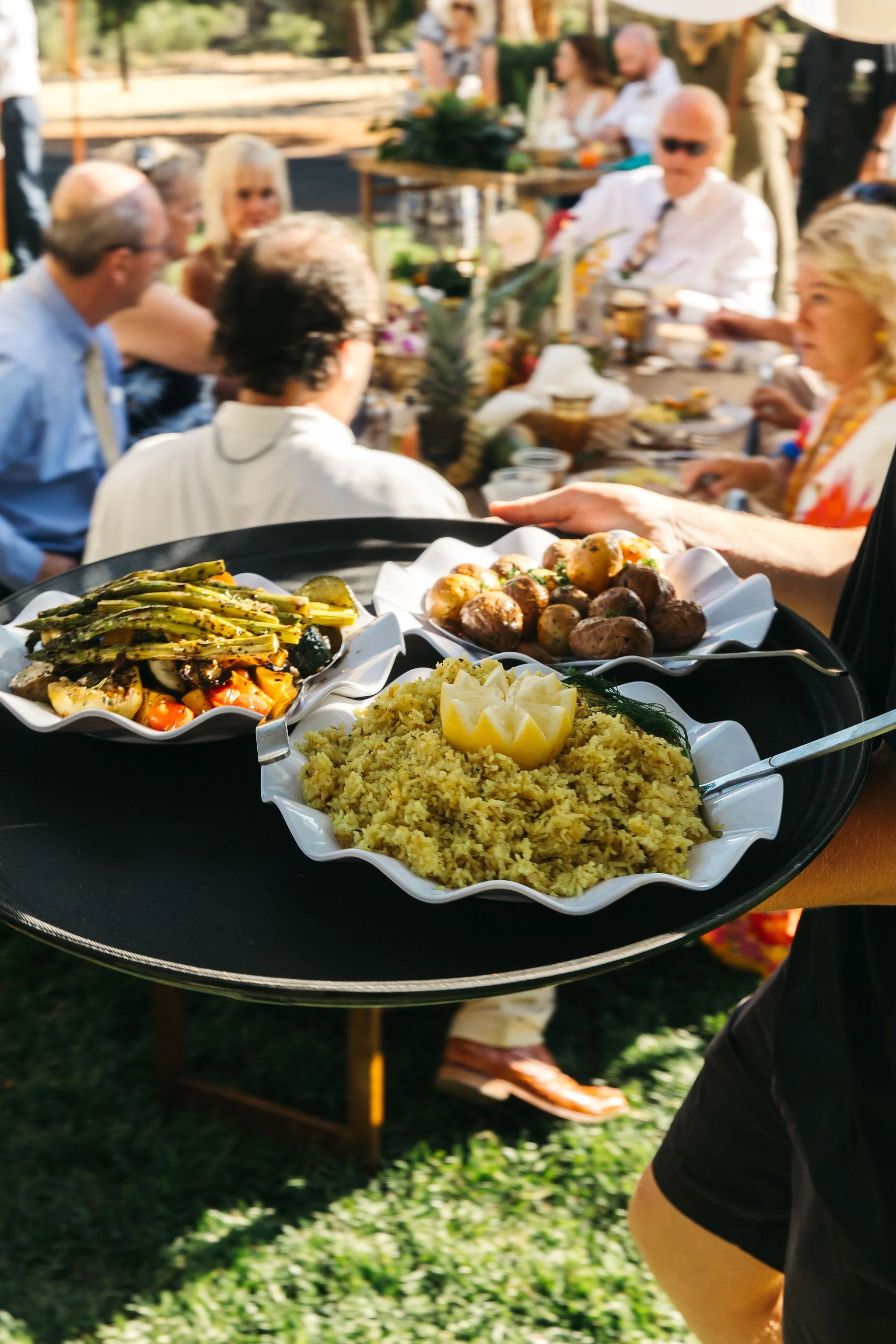 A person holding a tray with bowls of food, including roasted vegetables, mashed potatoes, and a grain dish, at an outdoor gathering with people dining at a long table in the background.