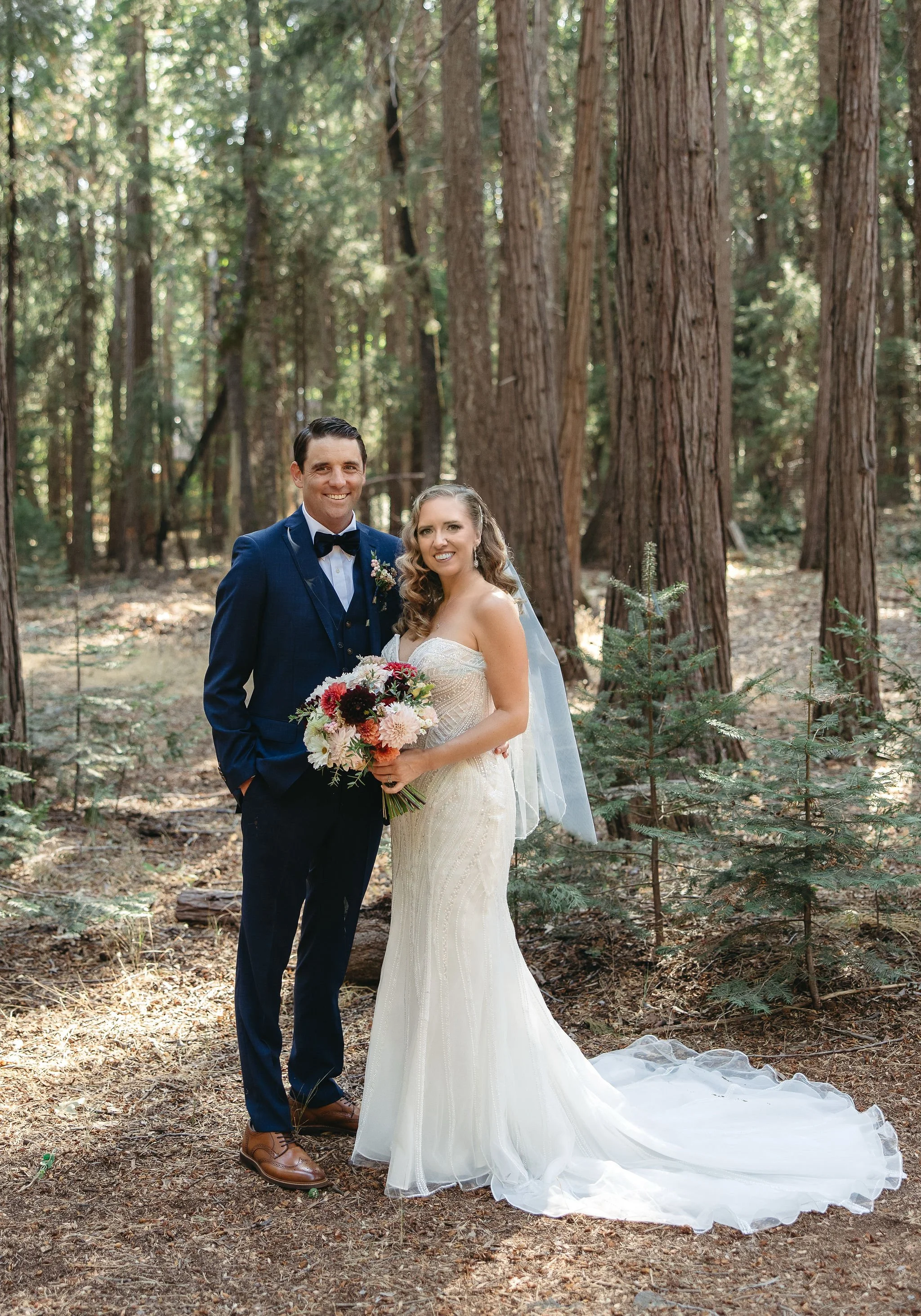 A bride and groom standing together in a forest with tall trees, smiling, with the bride holding a large bouquet of flowers, and both dressed in wedding attire.