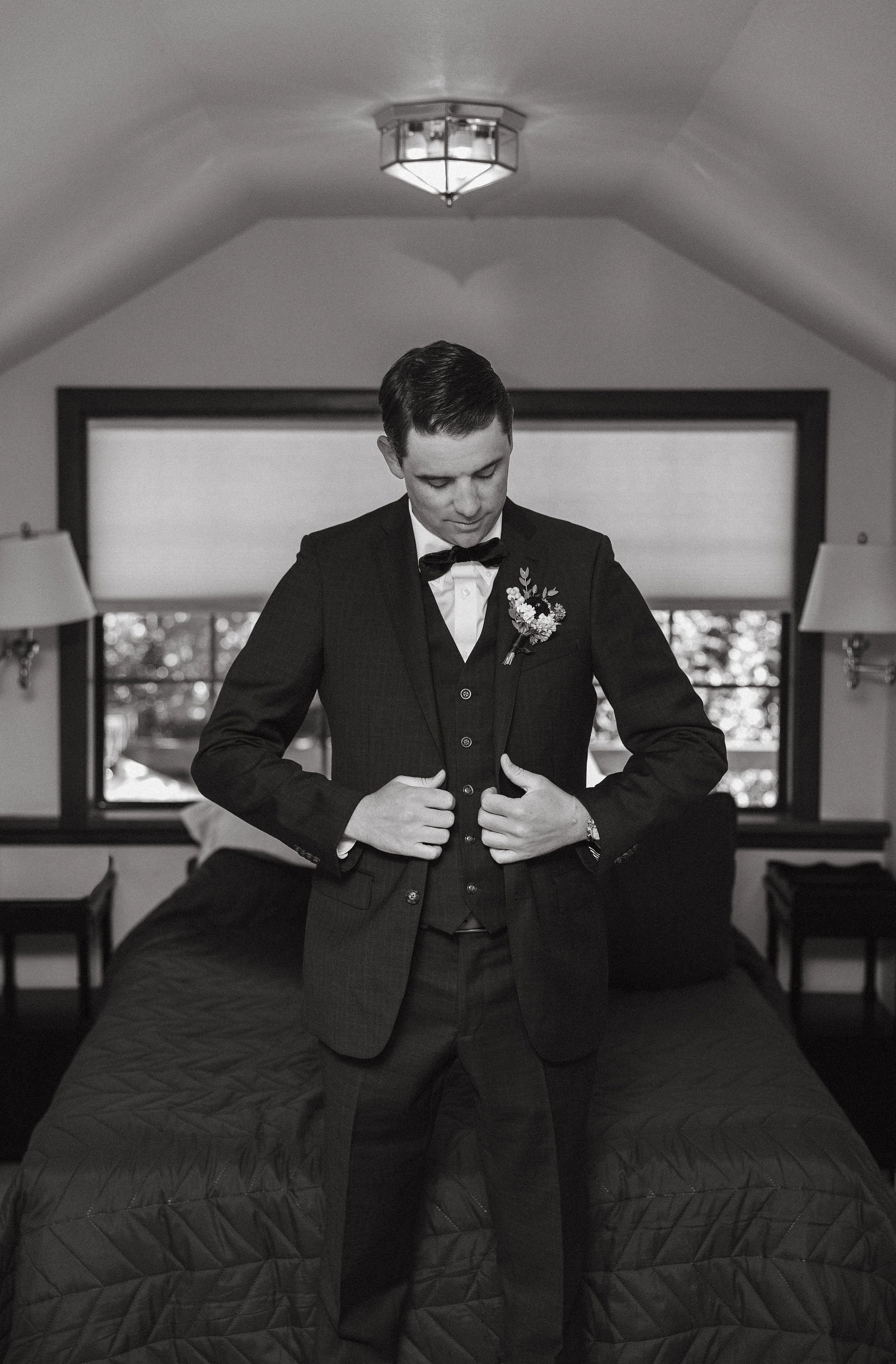 A man in a tuxedo with a boutonniere on his lapel standing on a bed, adjusting his jacket.