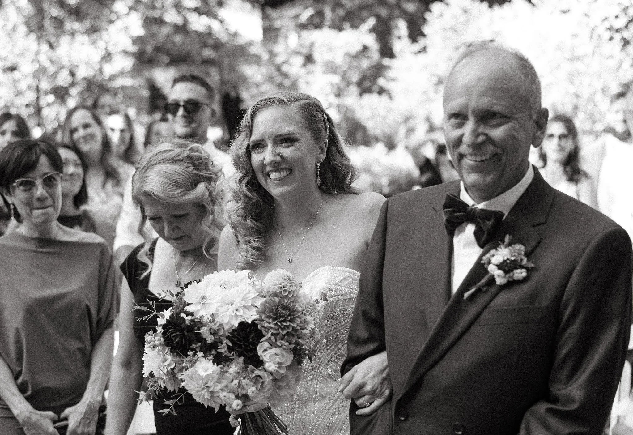 A bride holding a bouquet of flowers, smiling, walking down the aisle with an older man in a suit and bow tie, surrounded by guests at an outdoor wedding ceremony.