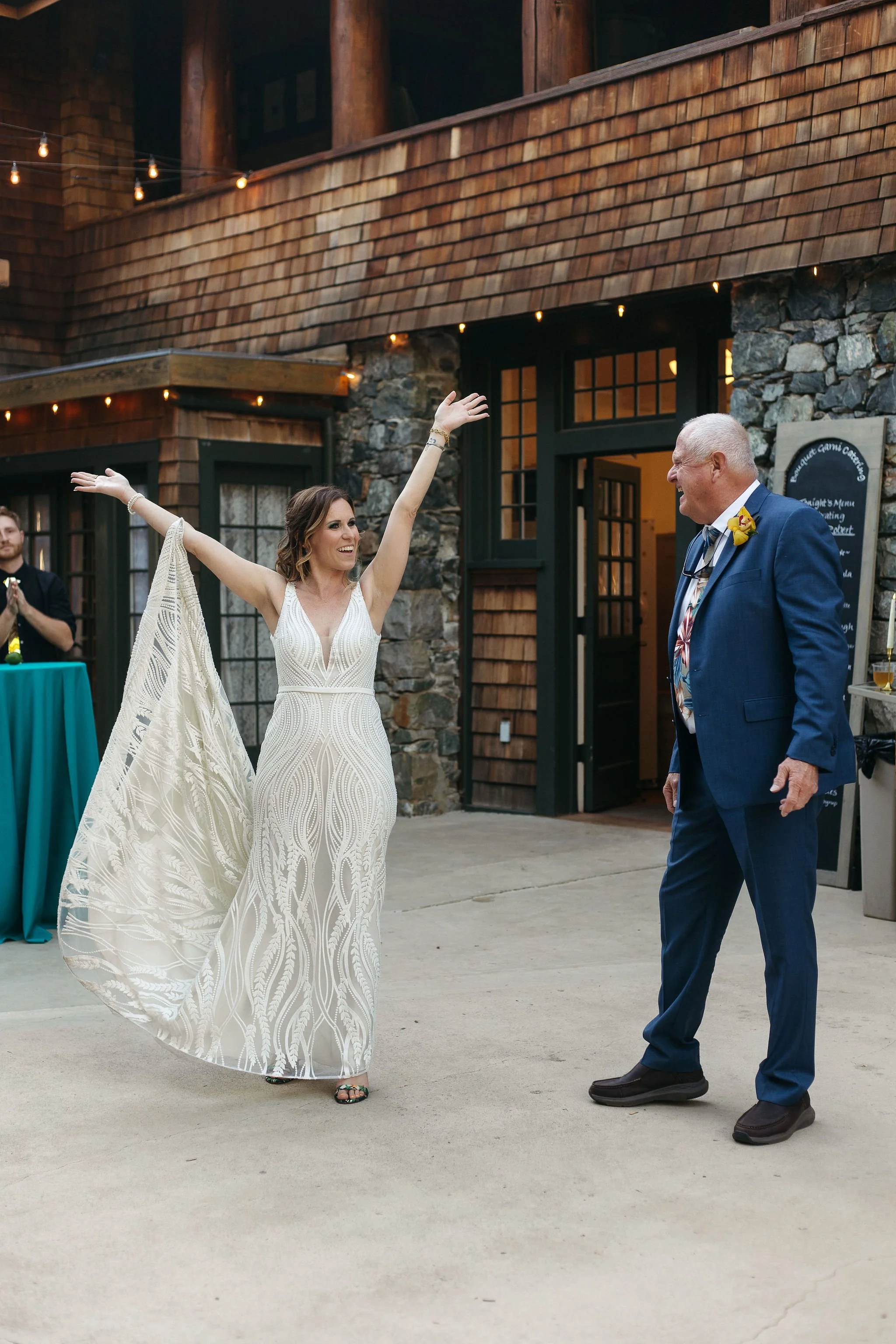 Woman in an elegant white dress with arms raised and smiling, facing a man in a blue suit with a yellow flower boutonniere, both enjoying a joyful moment at a celebration outside a rustic venue with stone and wood exterior.