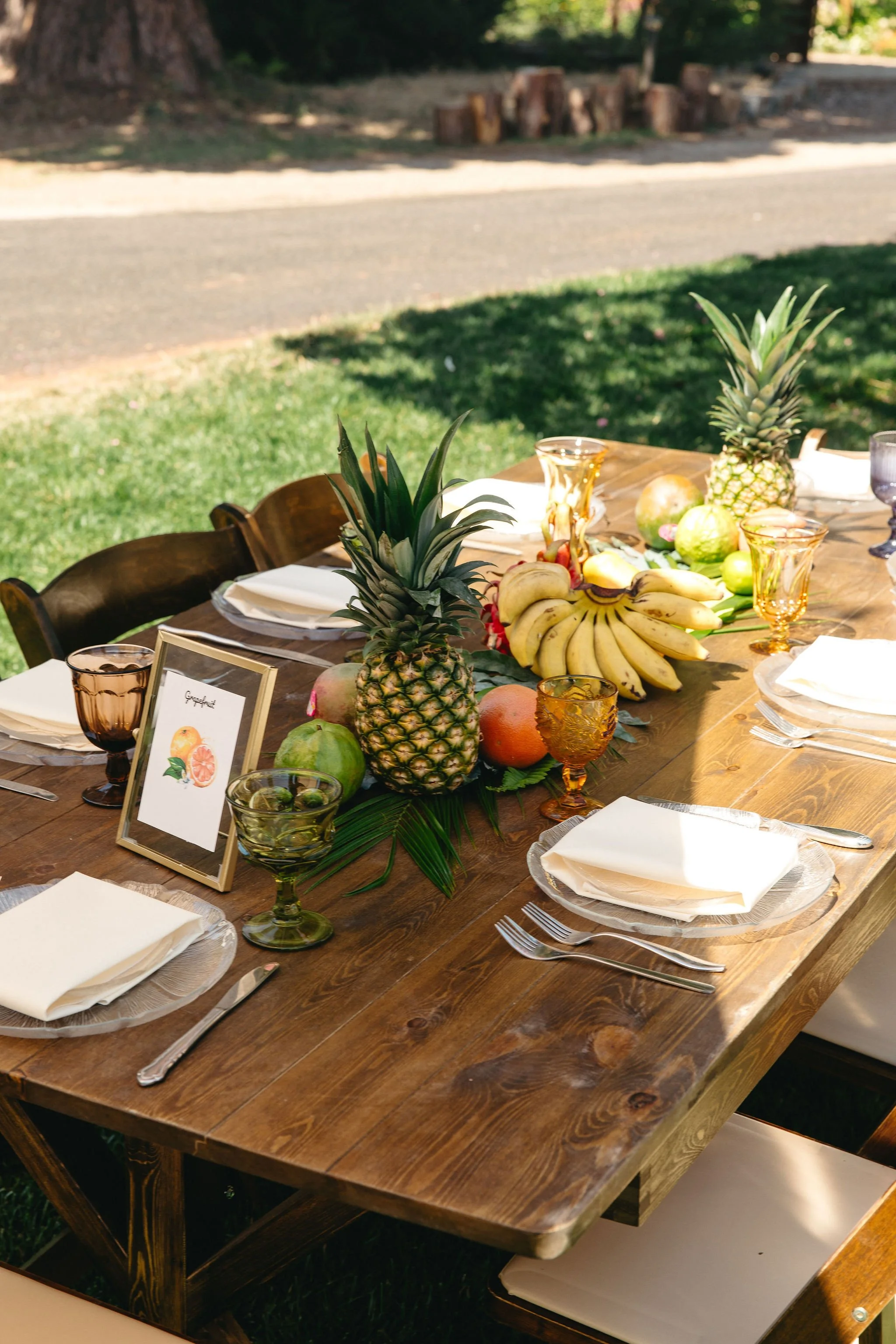 A wooden outdoor table set for a meal with white plates, folded napkins, silverware, and colorful glasses. The table has a centerpiece of pineapples, bananas, green apples, oranges, and other tropical fruits, with a small framed sign featuring an ill