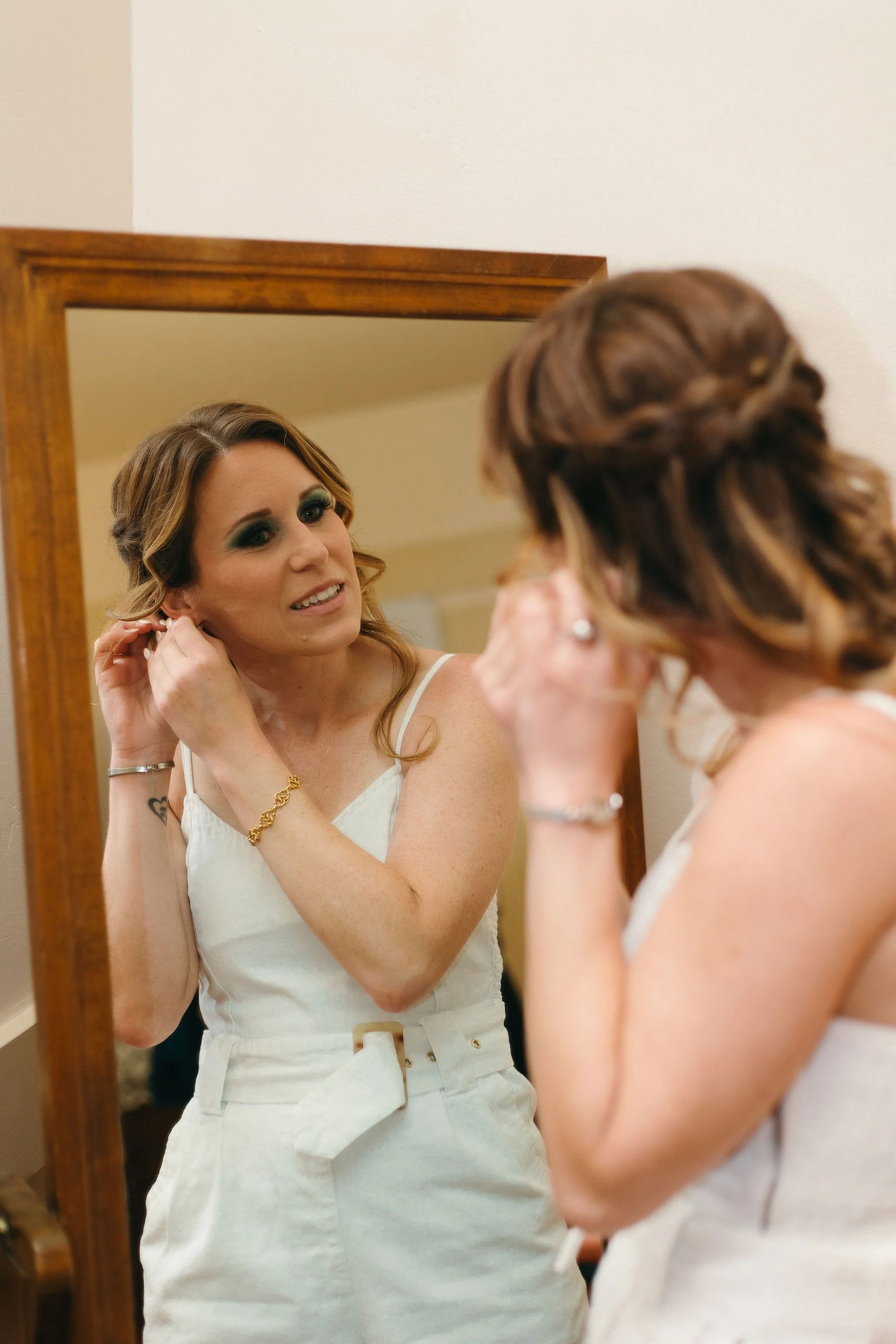 Woman with makeup and jewelry adjusting her earring while looking in a mirror.