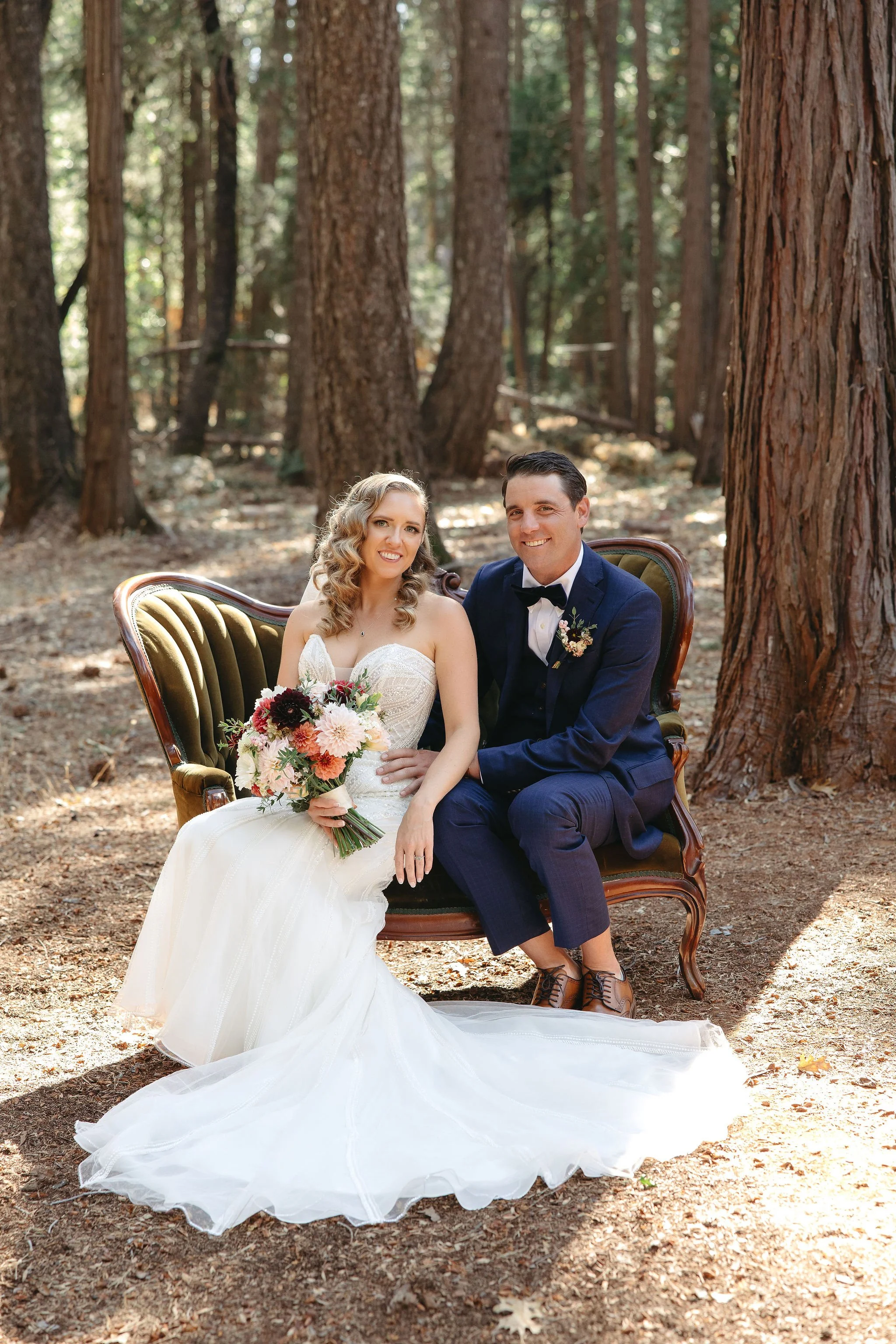 A bride and groom sitting on a vintage green velvet sofa in a forest clearing during their wedding photos. The bride wears a white strapless wedding dress and holds a bouquet of flowers. The groom wears a navy blue suit with a bow tie. They are smili