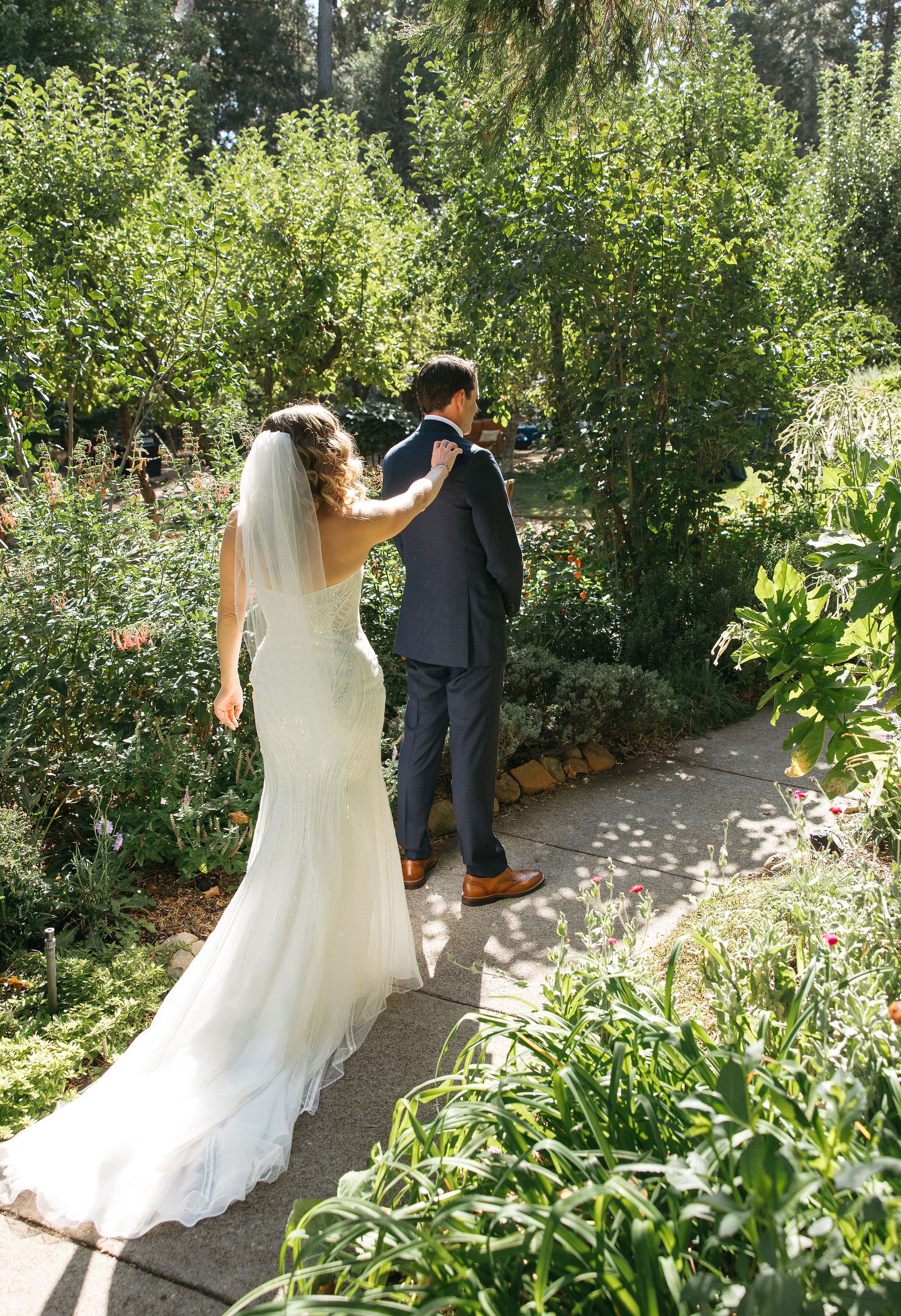 A bride and groom walk along a garden path, with the bride gently touching the groom's shoulder, during their wedding.