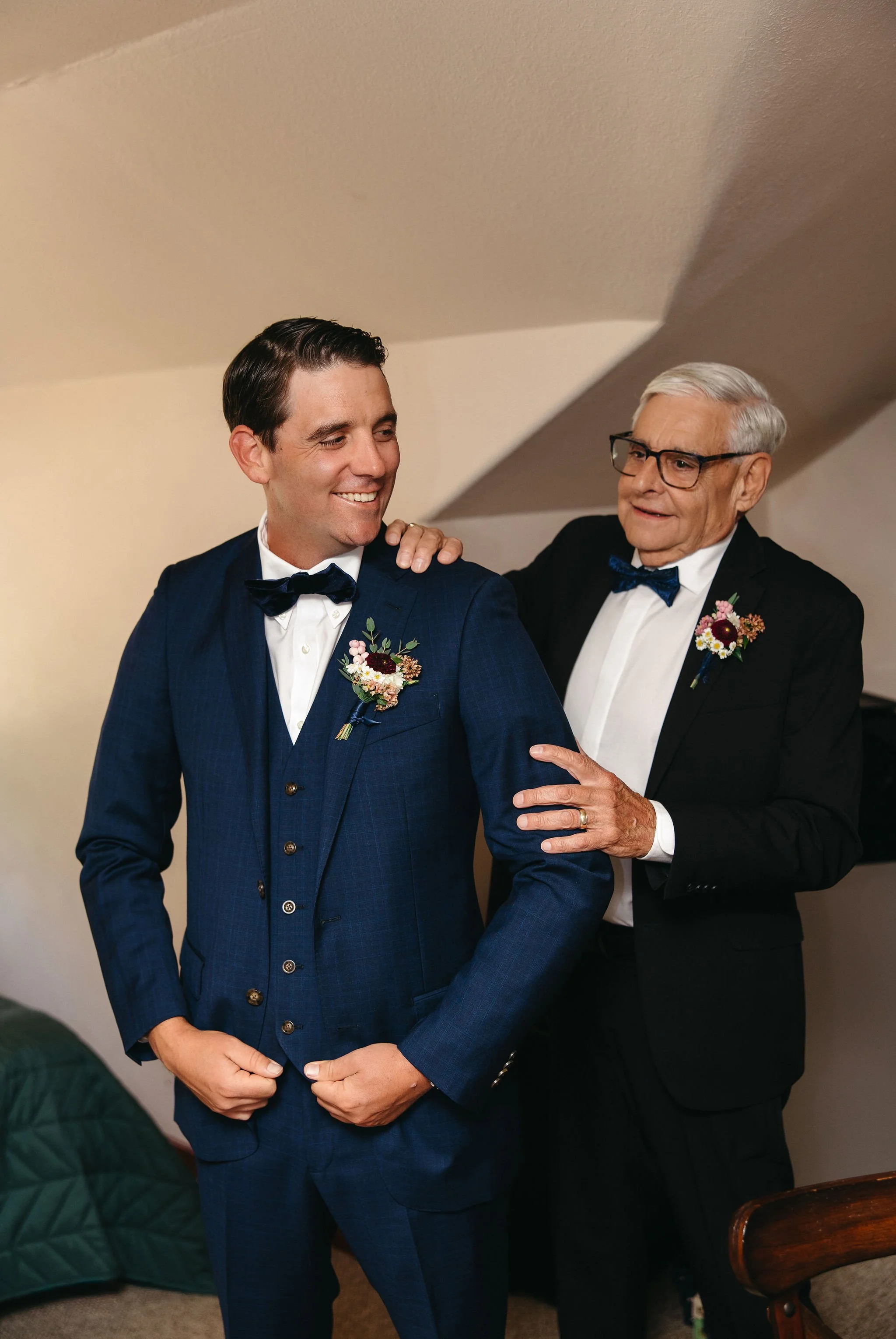 Older man with glasses helping a younger man adjust his suit, both dressed in tuxedos with boutonnières, in a warmly lit room.