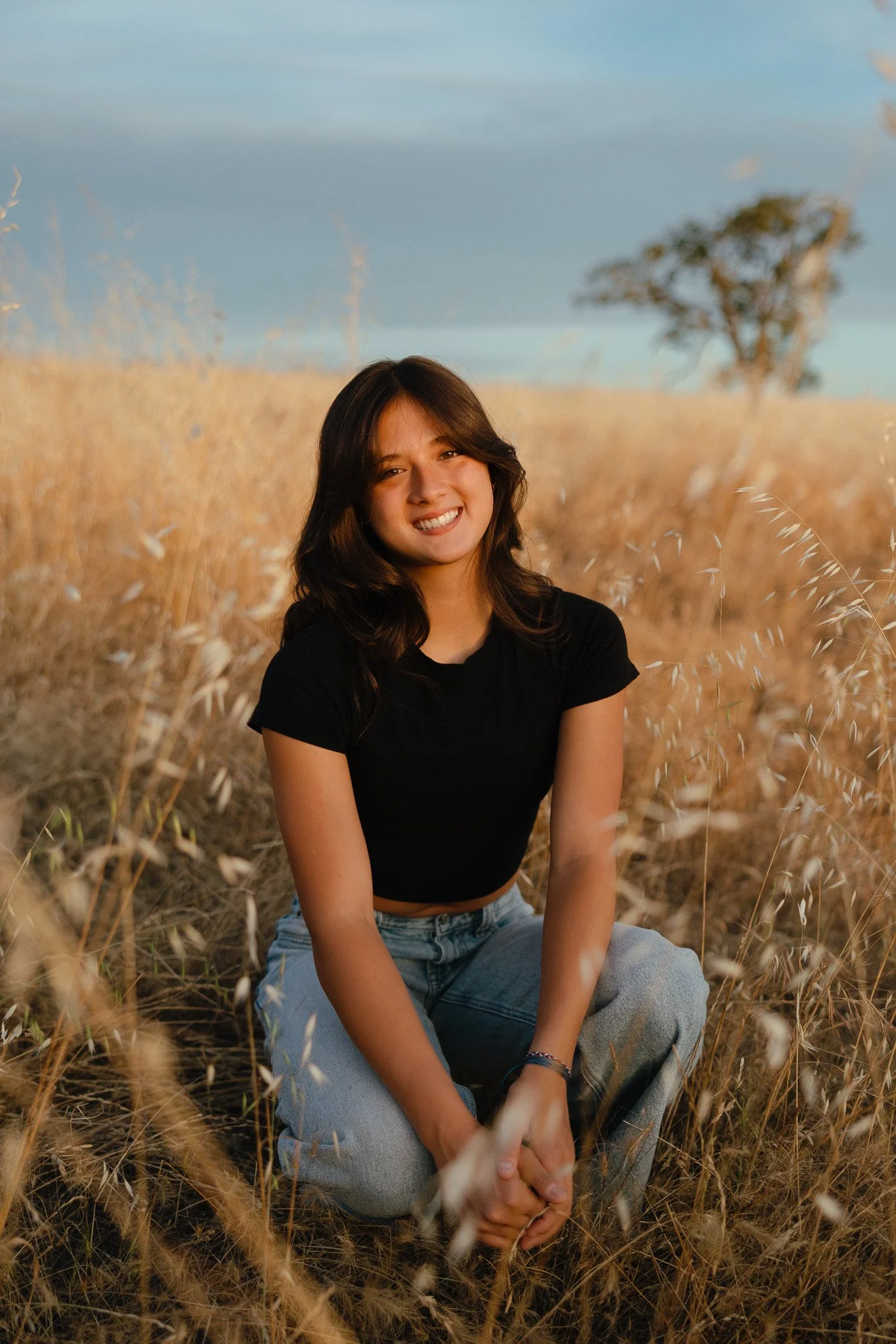 A young woman with brown hair, wearing a black t-shirt and jeans, sitting in a field of tall, dry grass during sunset, smiling at the camera.