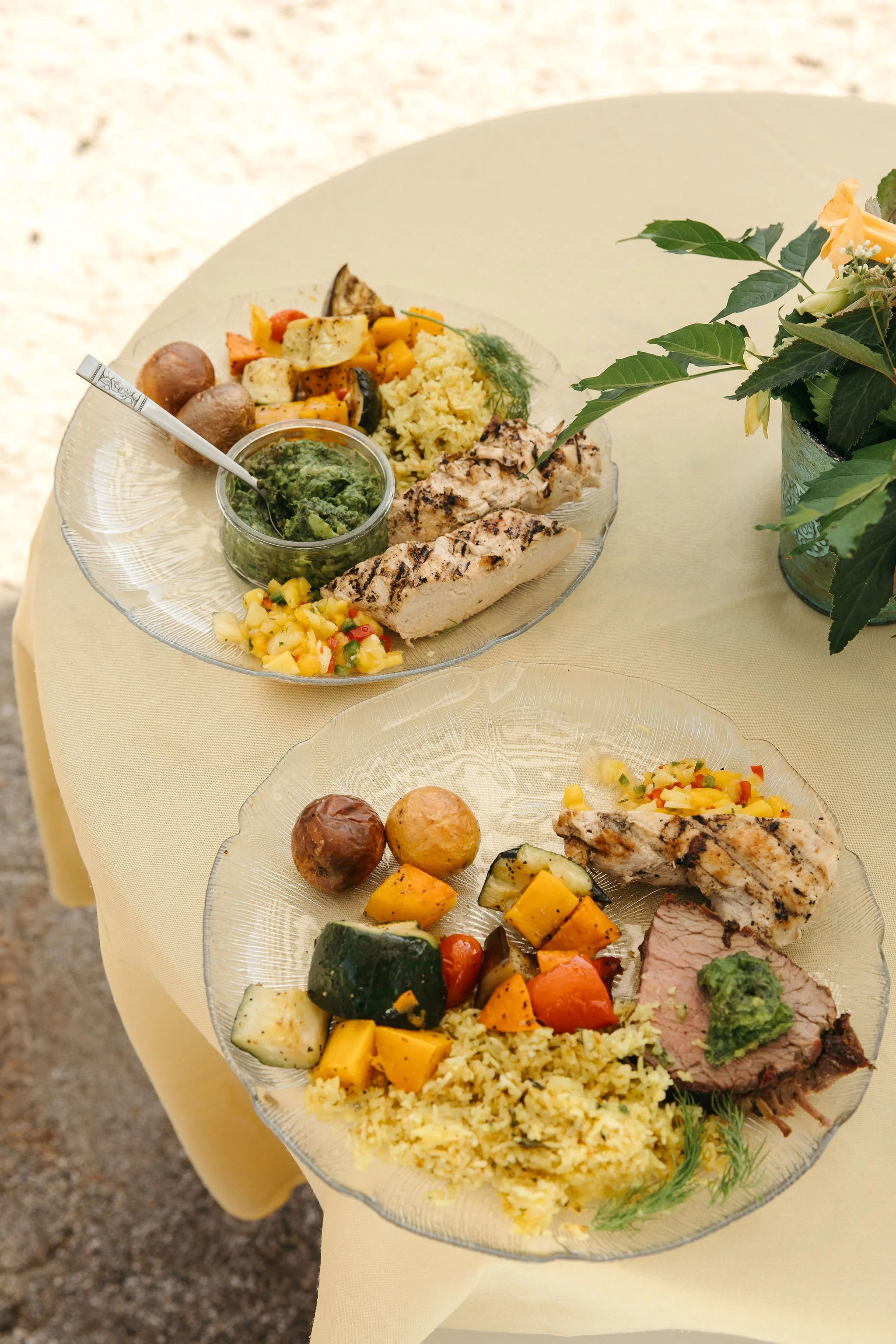 Two glass plates with grilled chicken, roasted vegetables, rice, and salad on a yellow tablecloth, with a potted plant nearby.