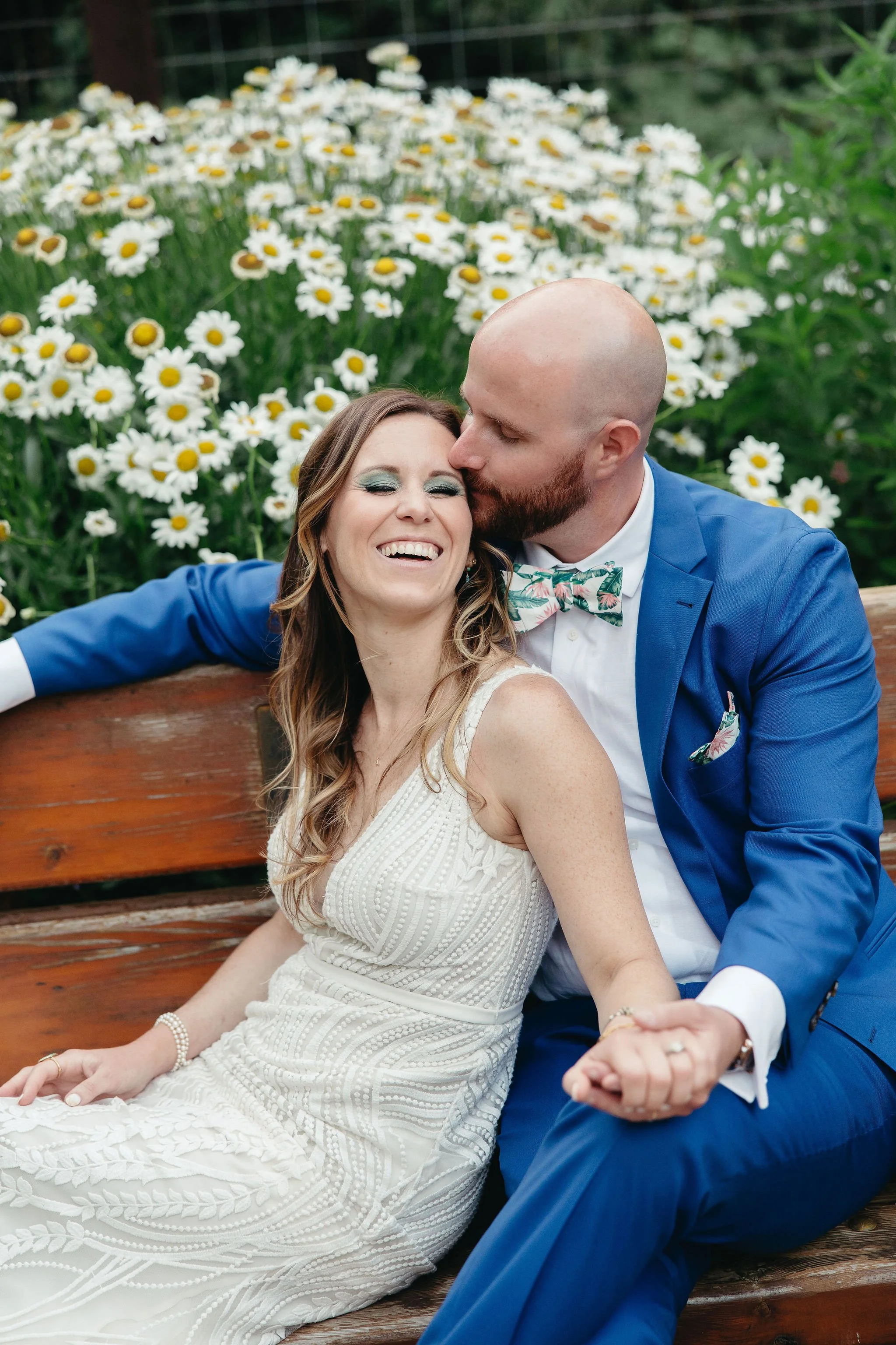 A joyful woman in a white dress and a man in a blue suit sit on a wooden bench, sharing a kiss with a background of white daisies.