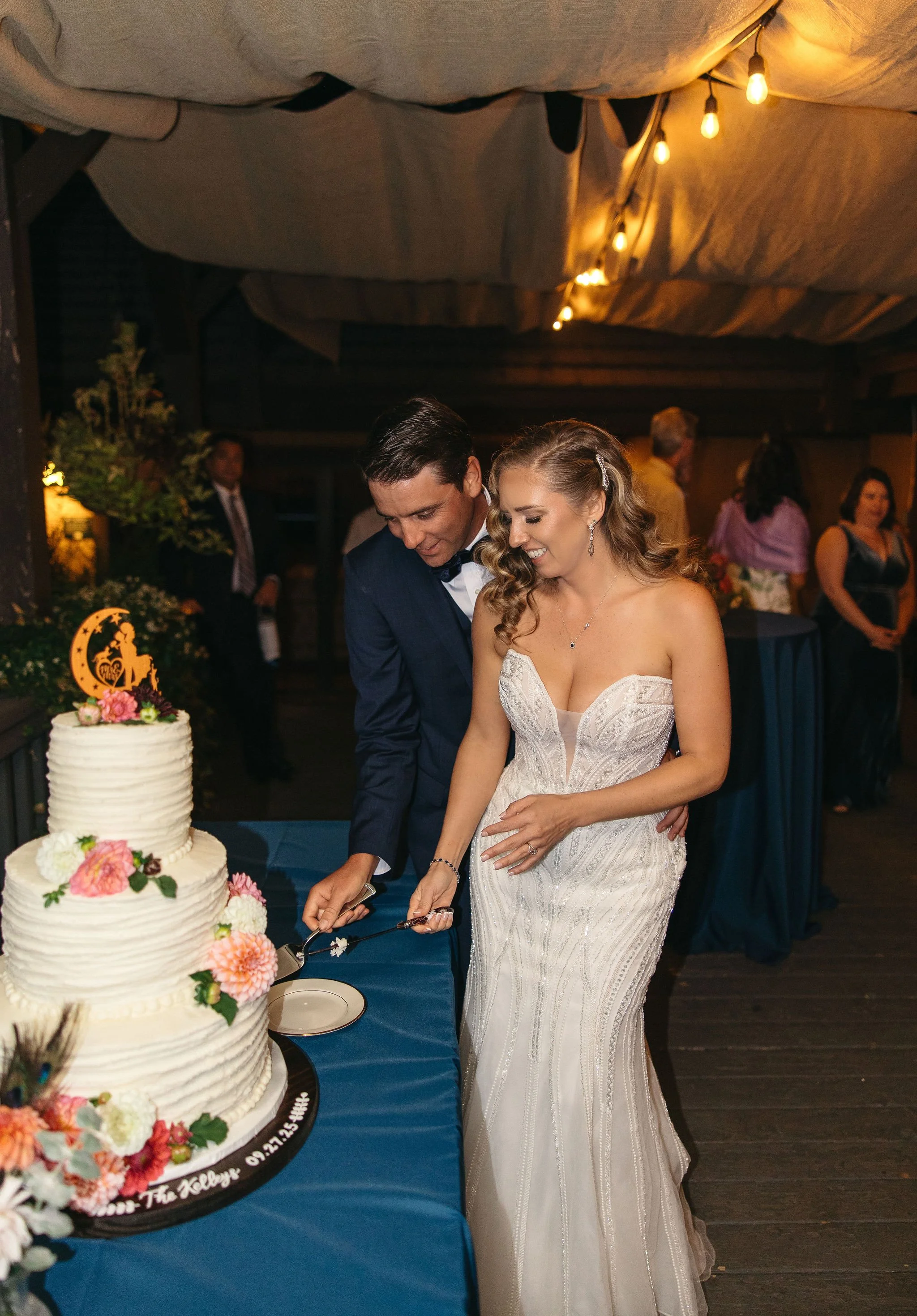 Bride and groom cutting a wedding cake at their reception, with wedding guests in the background.