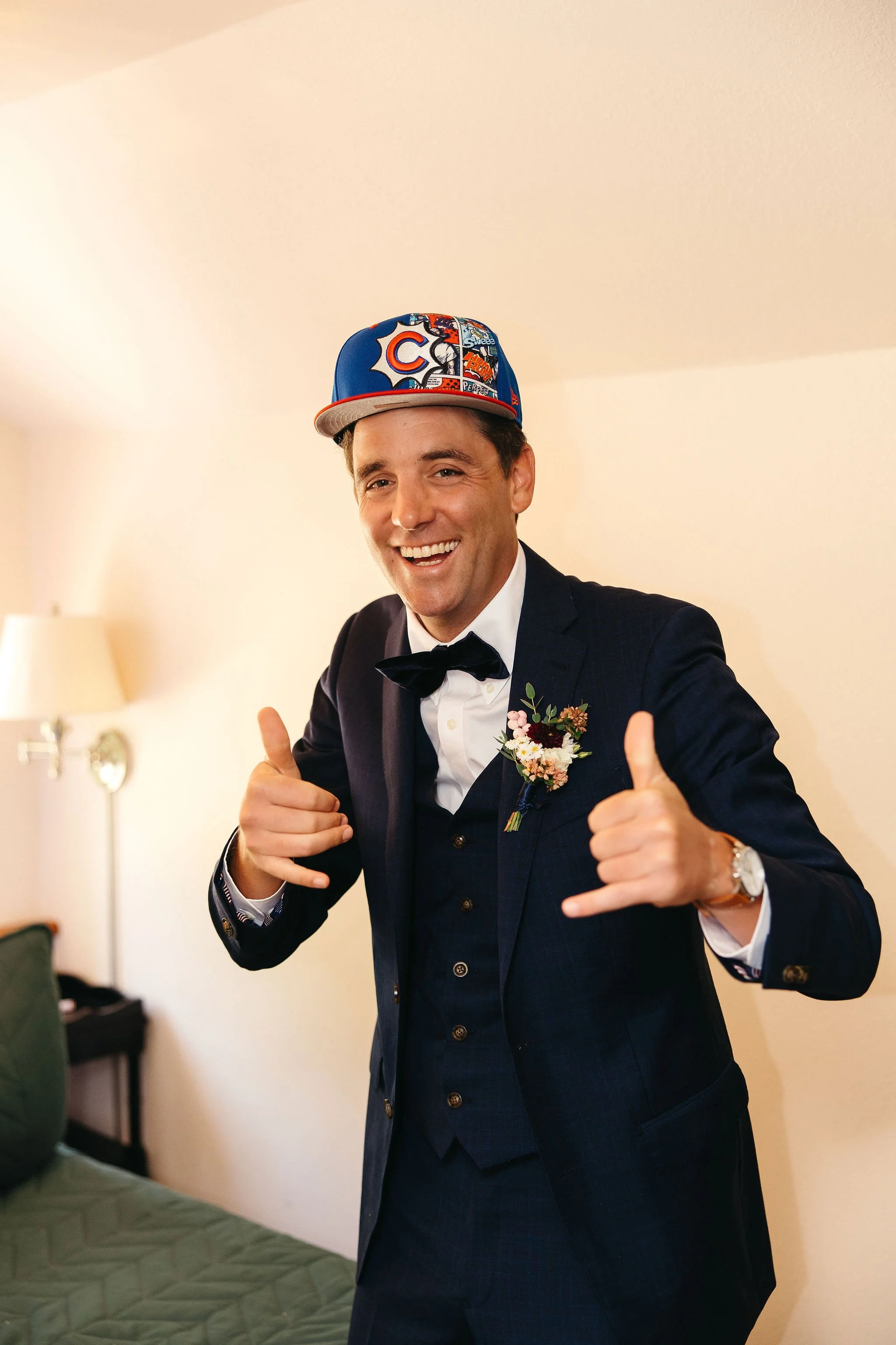 A man dressed in a suit with a flower boutonniere, smiling and making a shaka sign with both hands, wearing a Chicago Cubs cap.