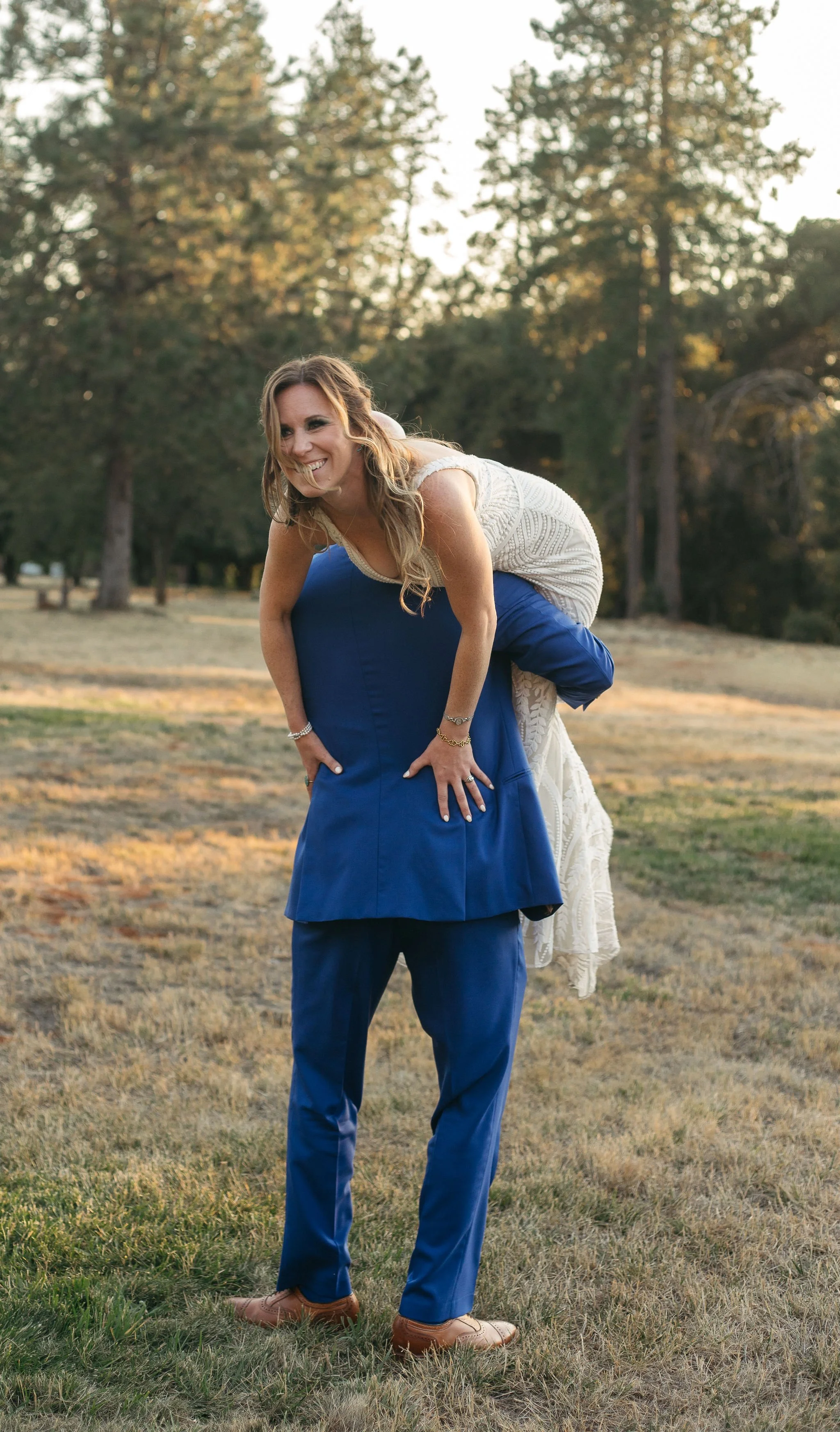 A woman in a white dress is giving a piggyback ride to a woman in a blue suit outdoors during daytime.