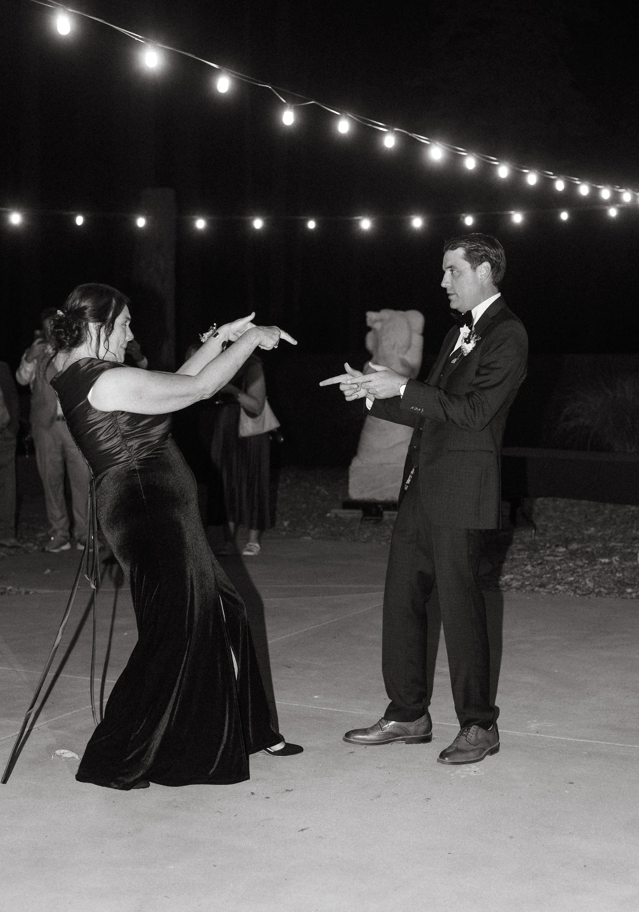 A bride and groom dancing at a wedding reception outdoors at night, with string lights overhead and guests in the background.