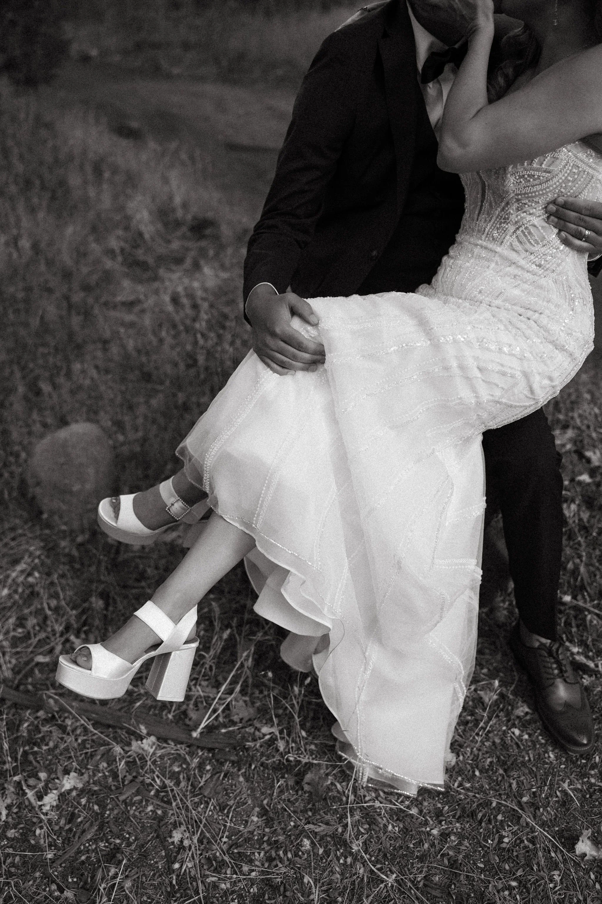 A bride in a wedding gown being carried by a groom in a suit, outdoors on grass.
