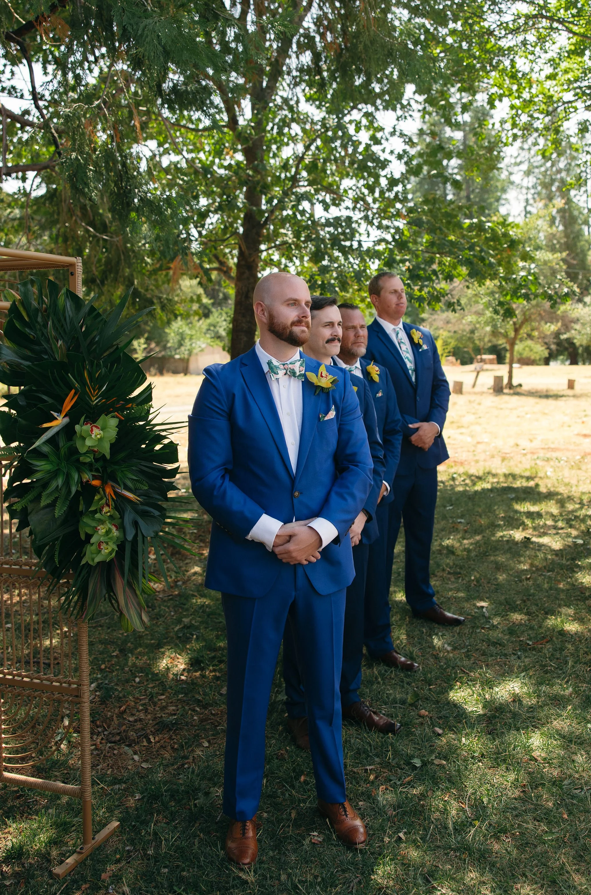 Four men dressed in blue suits with floral bow ties and yellow boutonnieres stand outdoors at a wedding ceremony, lined up near a lush green tree and decorated with tropical flowers and foliage.