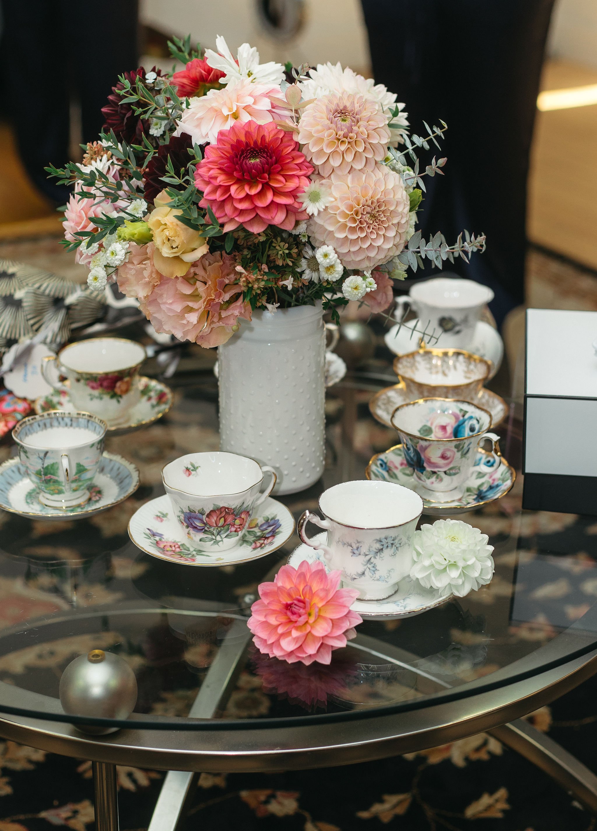 A glass table with a large bouquet of pink, white, and red flowers in a white vase, surrounded by vintage teacups and saucers, some decorated with floral patterns. There is also a single pink flower and a pearl-like object on the table.