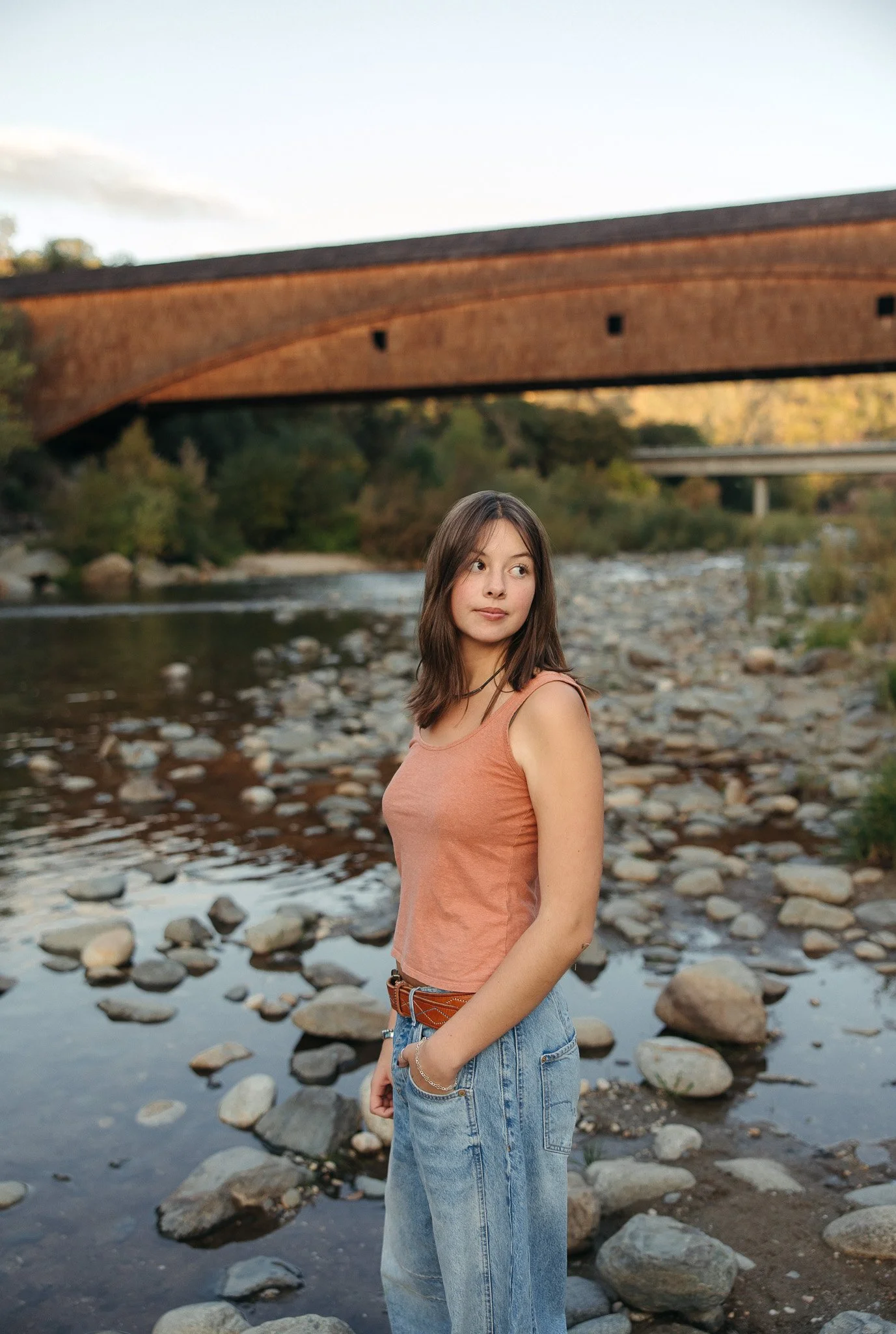 A young woman with long brown hair wearing a peach tank top and high-waisted jeans stands near a rocky riverbank with a bridge overhead, trees in the background, during sunset.