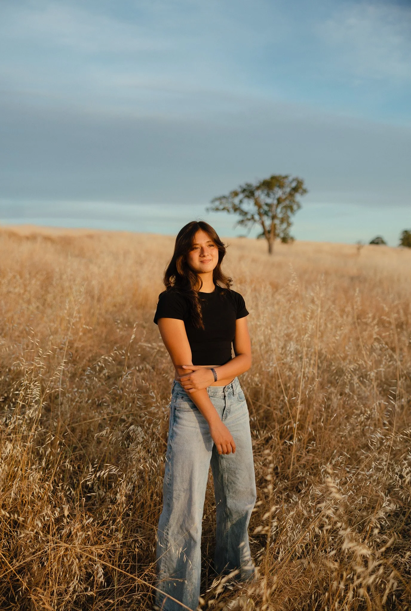 A young woman with long dark hair wearing a black t-shirt and light blue jeans standing in a field of dry golden grass with a solitary tree in the background under a partly cloudy sky.