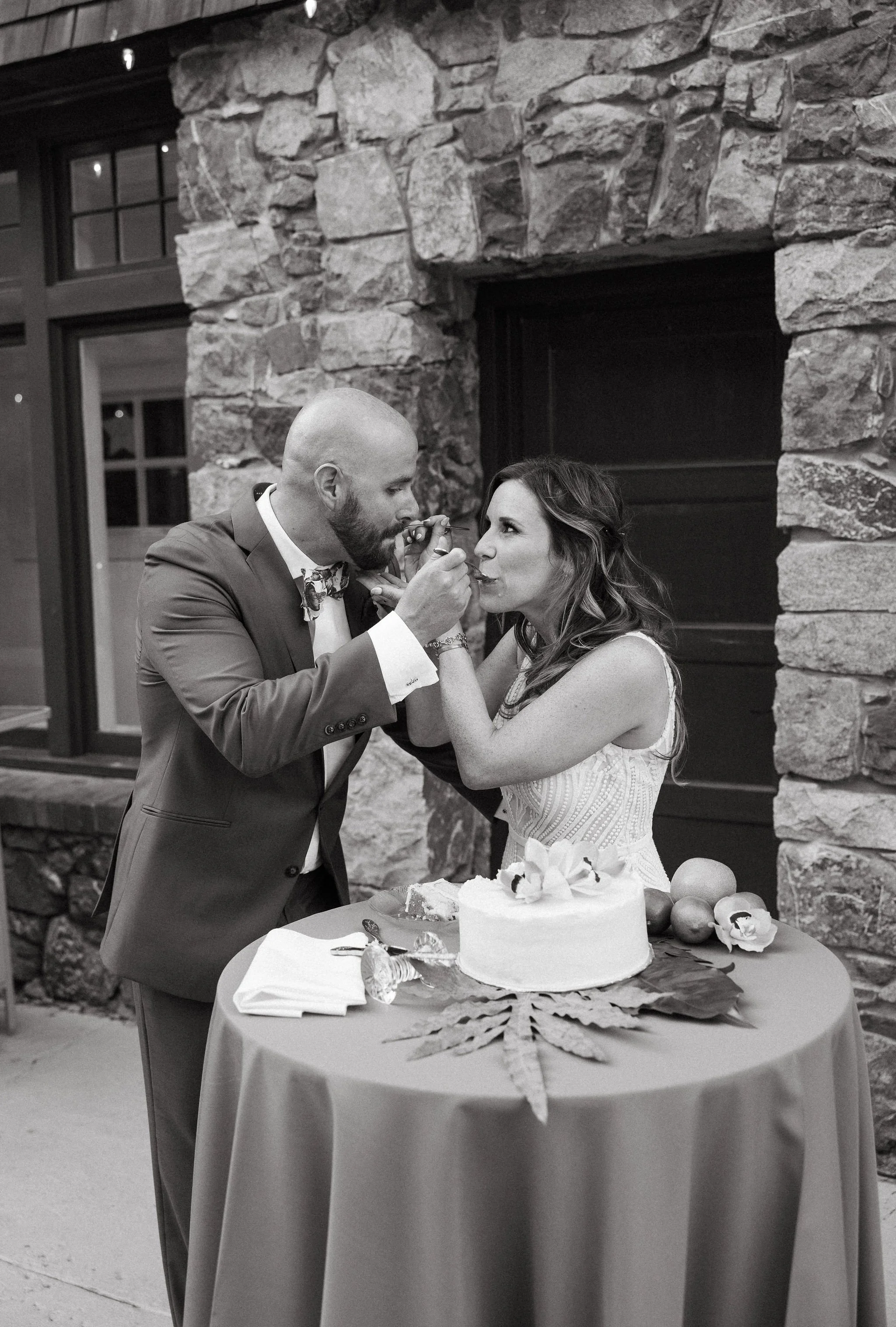 A man and woman sharing a piece of cake at a wedding reception, with a wedding cake and fruit on the table.