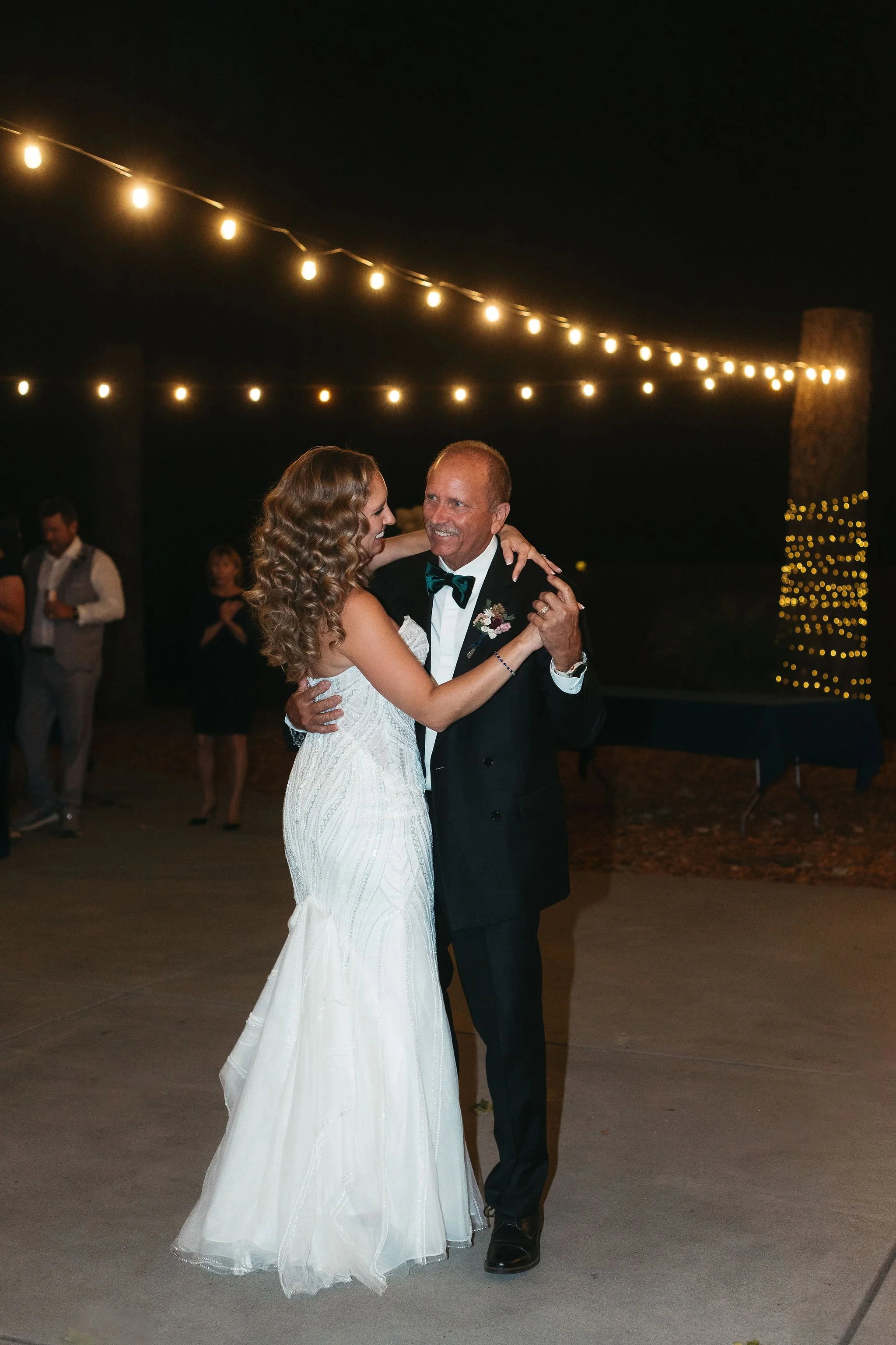 A bride and an older man, likely her father, dancing at a wedding reception outdoors at night, under string lights with some people in the background.