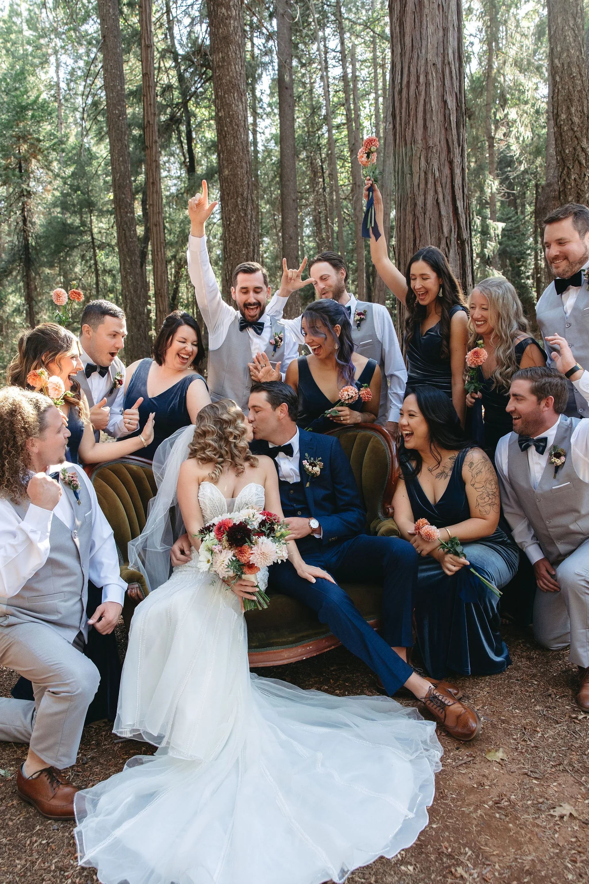 A wedding celebration in a forest with a bride and groom kissing on a vintage sofa surrounded by friends, some cheering, some holding flowers, all smiling and joyful.