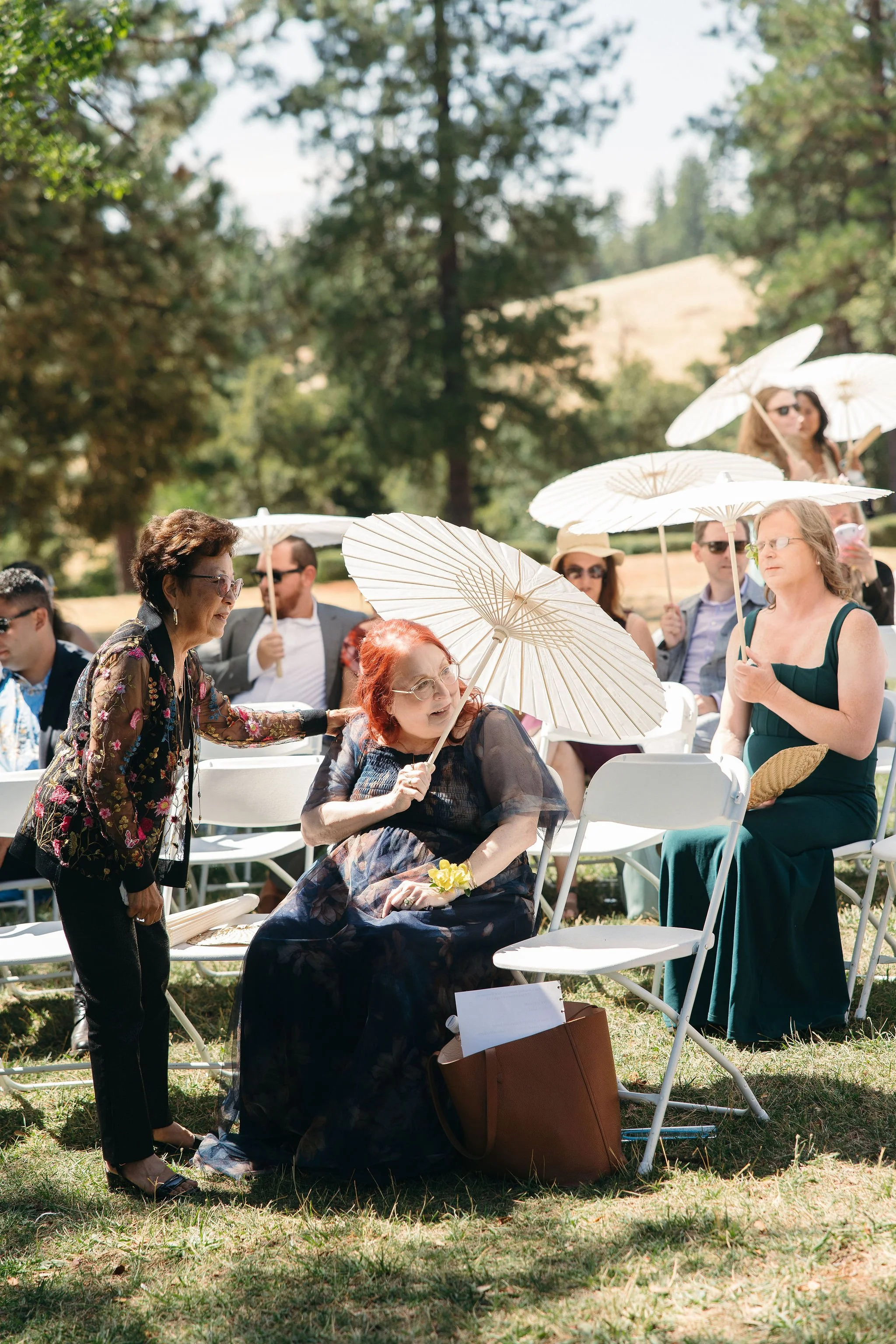 People sitting outdoors at a sunny event, holding white parasols, with a woman in a dark dress sitting in a chair with a parasol, and another woman standing beside her.