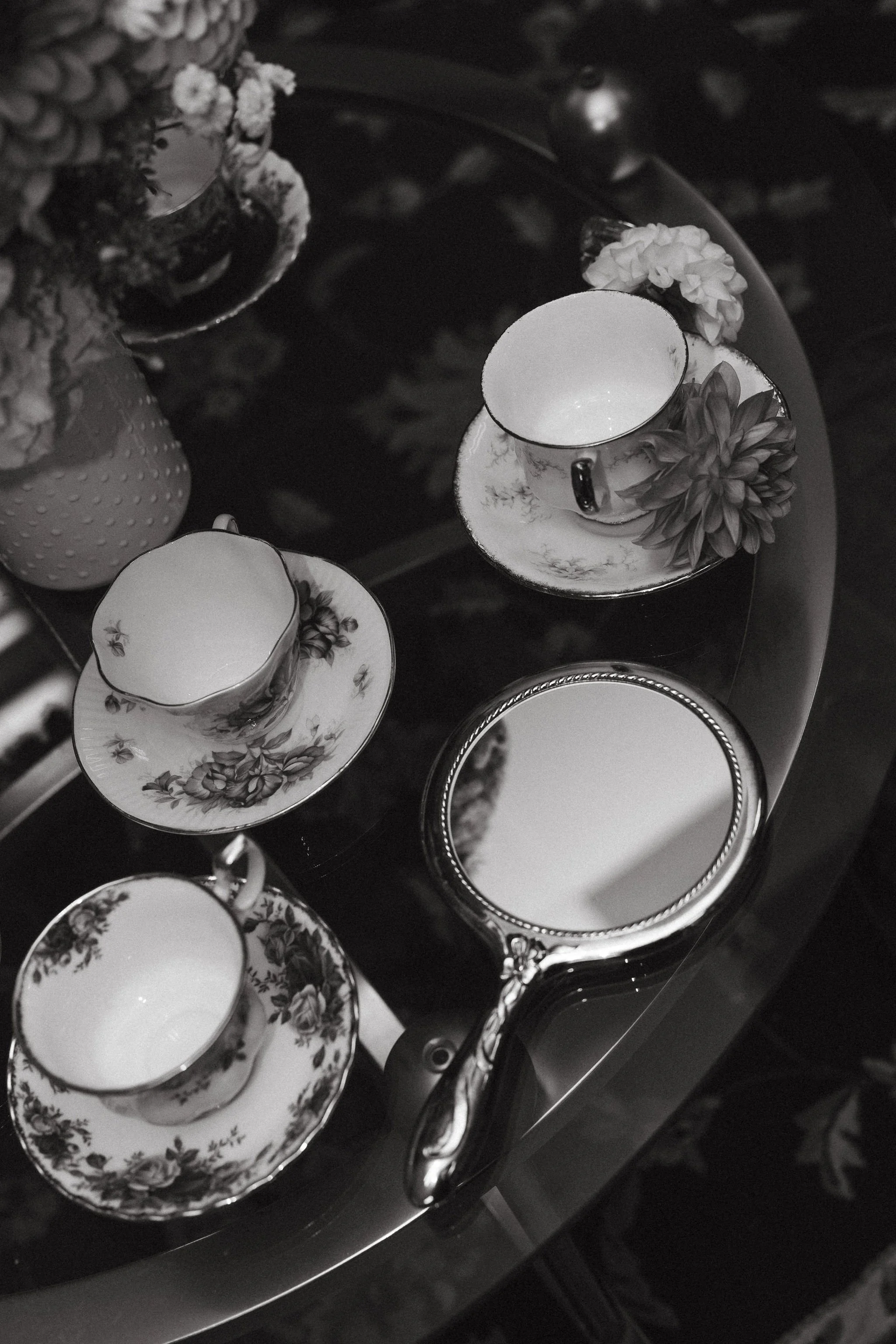 Three floral-patterned teacups and saucers on a table, with a round mirror and a flower arrangement nearby.