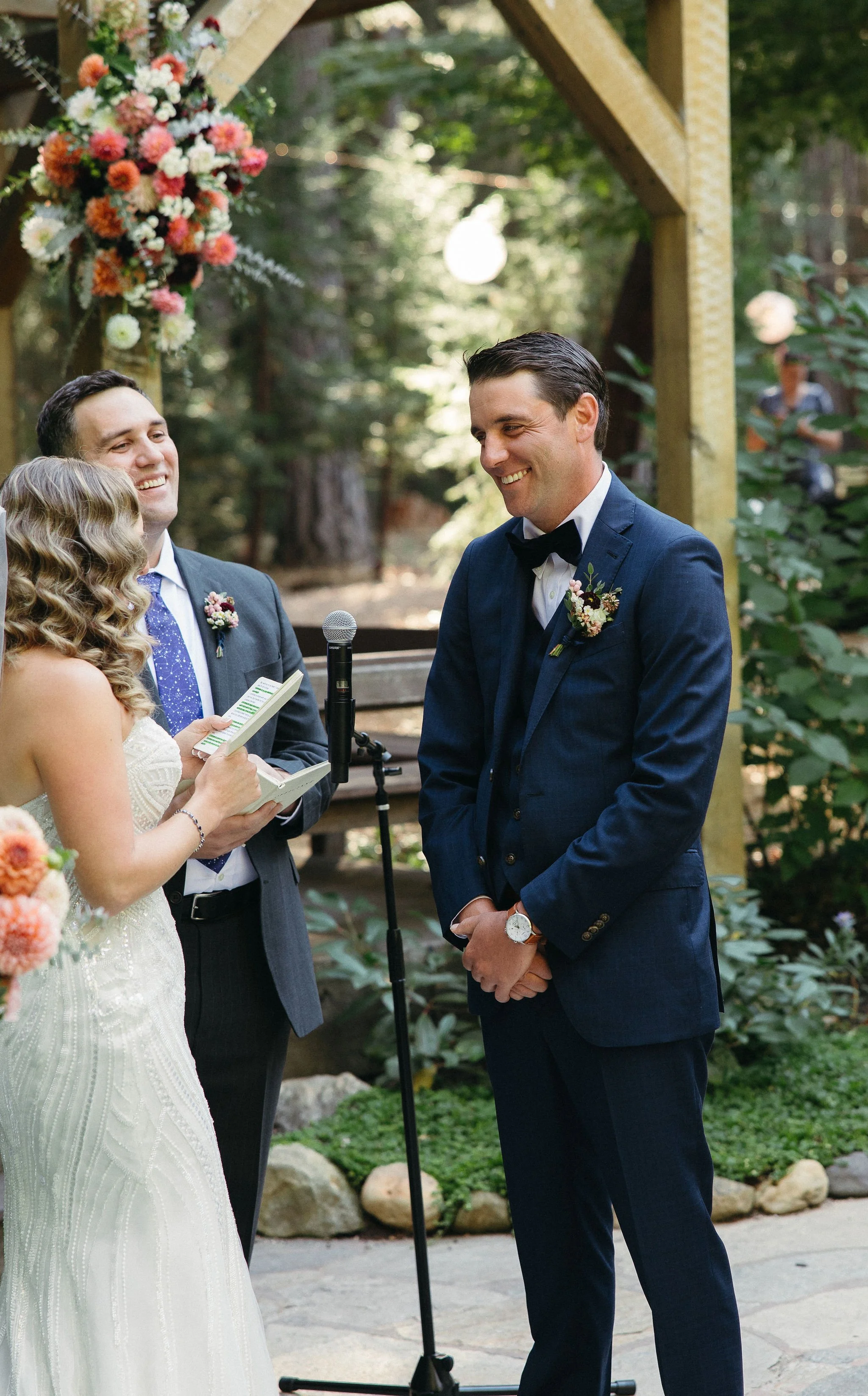 A wedding ceremony outdoors with the bride, groom, and officiant. The bride is holding papers, smiling, and wearing a white gown; the groom is in a navy suit with a boutonniere, smiling with clasped hands. The officiant is man in a suit holding notes