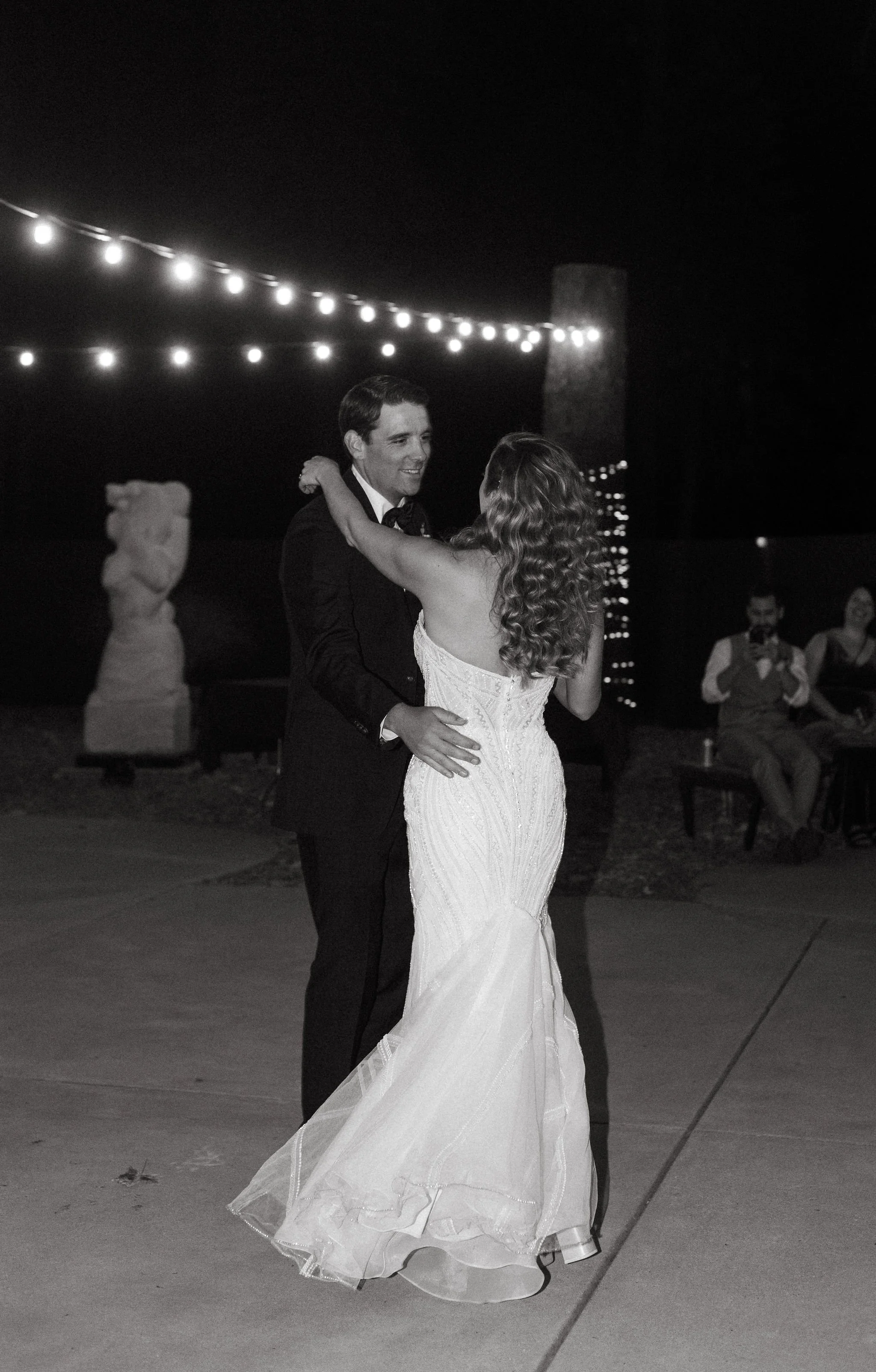 A black and white photo of a bride and groom dancing at night under outdoor string lights. The bride is in a strapless wedding gown with curly hair, and the groom is in a black tuxedo. There are statues and seated people in the background.