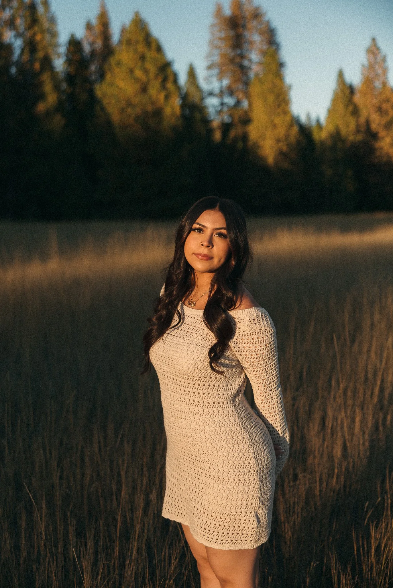 A young woman with long dark hair wearing a white knitted dress standing in a field of tall grass at sunset with a forest of pine trees in the background.