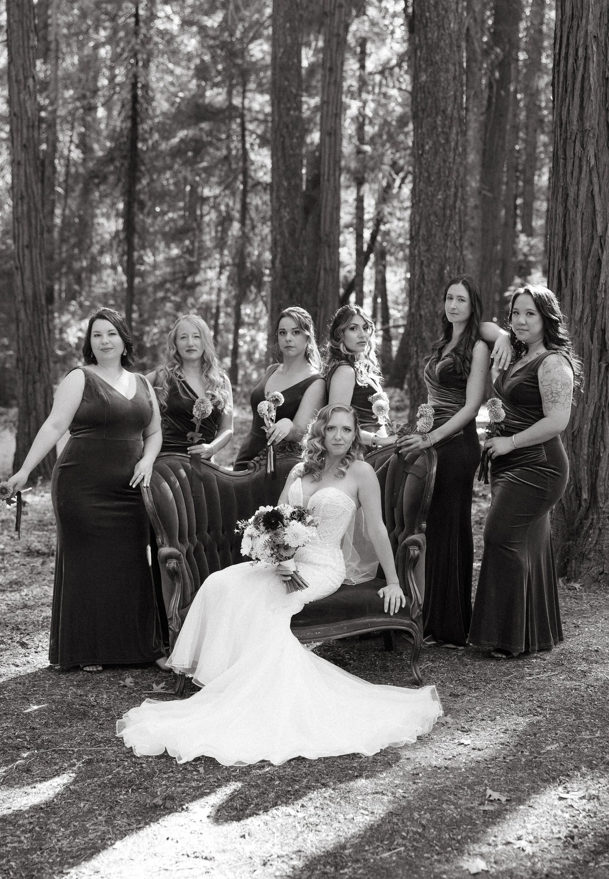 A bride and six bridesmaids in a forest, black and white photo. The bride is seated on a vintage sofa, holding a bouquet, wearing a strapless wedding gown. The bridesmaids are in long dark dresses, holding flowers, surrounding her.