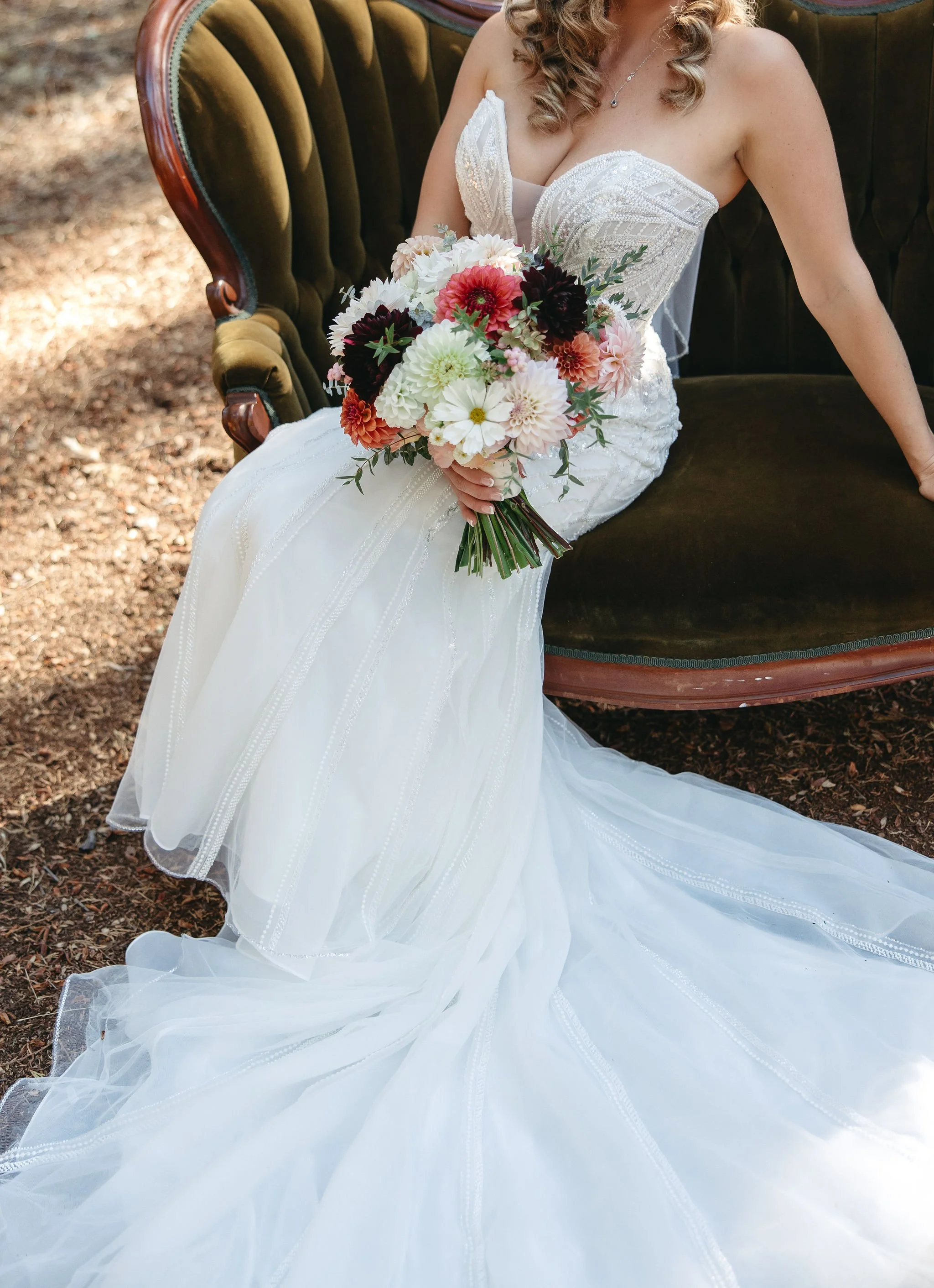 A bride sitting on a vintage sofa outdoors, holding a bouquet of mixed flowers. She wears a white wedding gown with intricate detailing, and her hair is styled in curls.