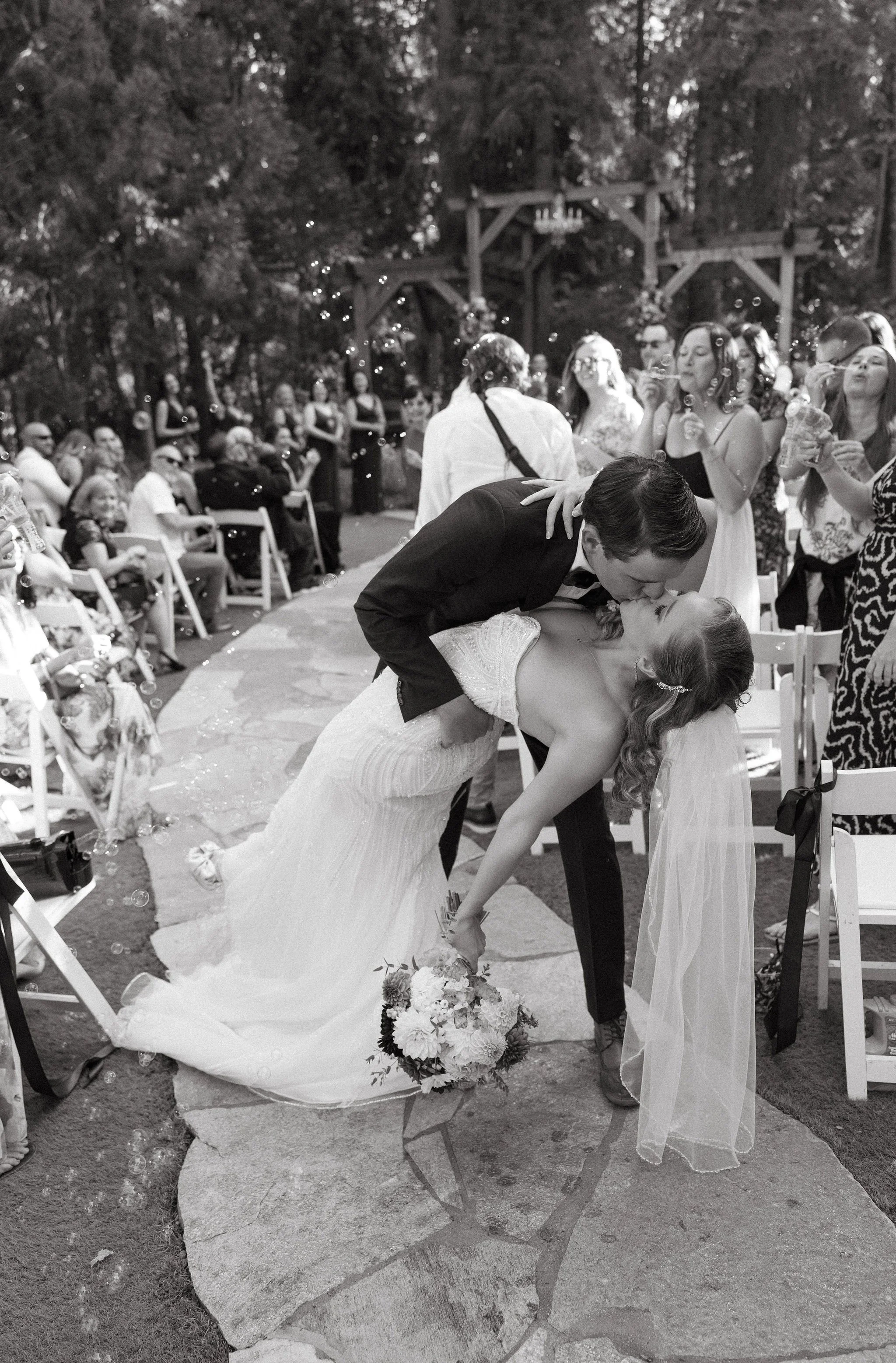 A wedding scene where the groom is kissing the bride while she leans back, holding a bouquet of flowers. They are surrounded by guests, with some sitting and others standing, in an outdoor setting with trees and decorations.