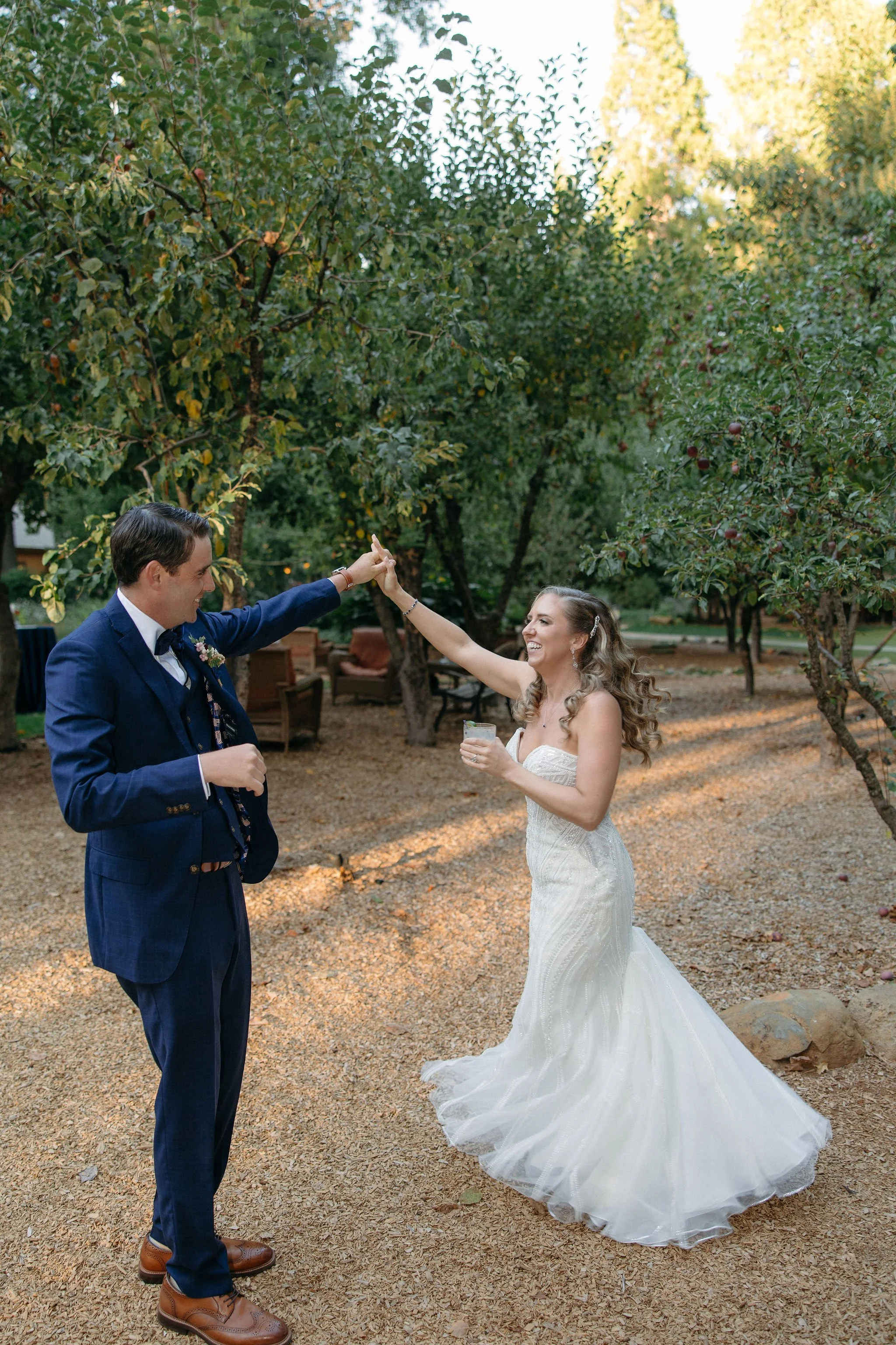 A bride and groom dancing outside under trees, with the groom wearing a navy suit and the bride in a white wedding gown holding a drink, smiling and touching hands.