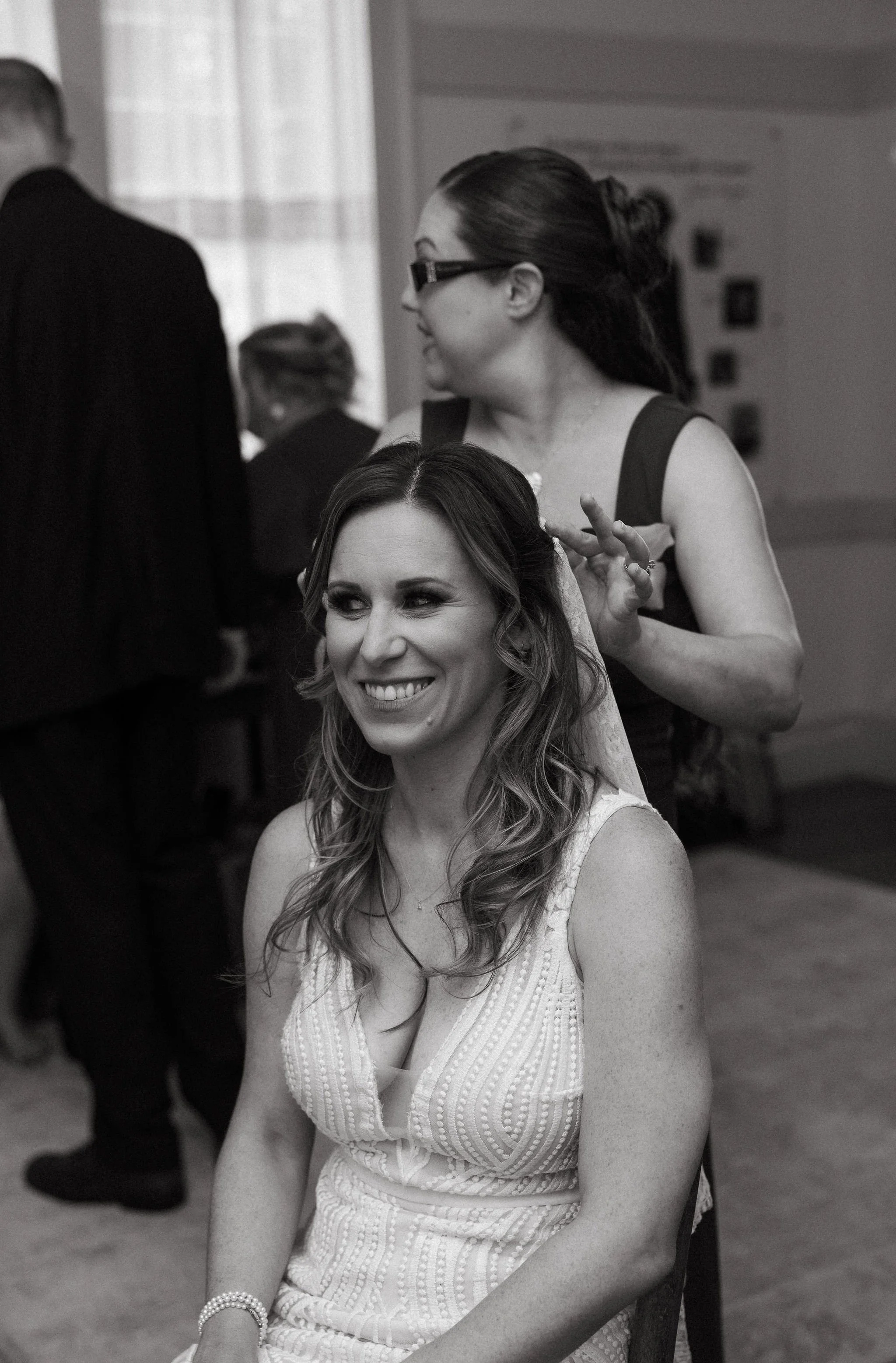 A woman with long wavy hair smiling while sitting down, having her hair styled by a woman standing behind her.